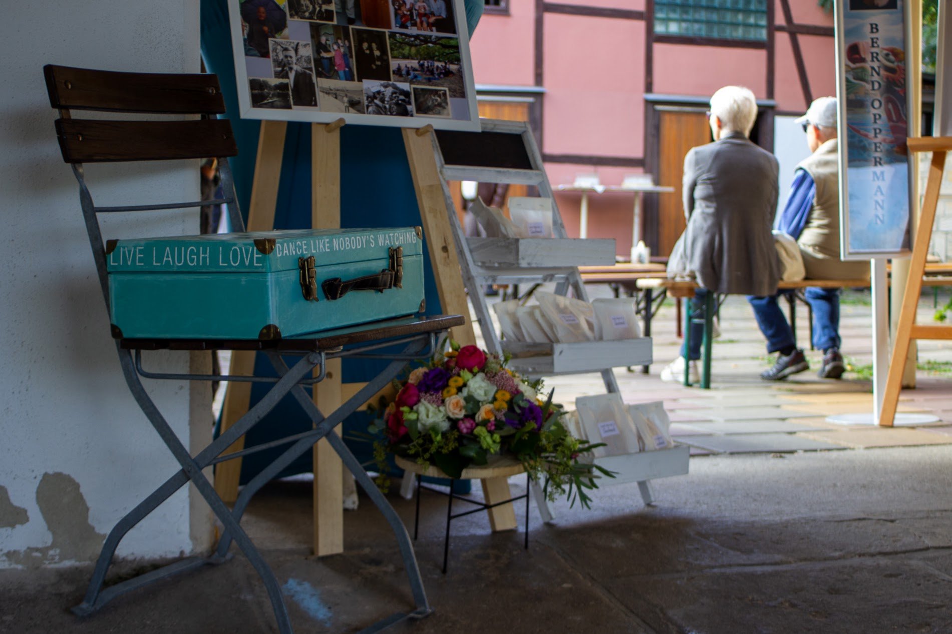 Ein Tisch mit einem blauen Koffer, einer Blumenvase und einem Fotowand im Vordergrund, im Hintergrund sitzen zwei ältere Männer auf einer Bank und unterhalten sich in einem kleinen Außenbereich.