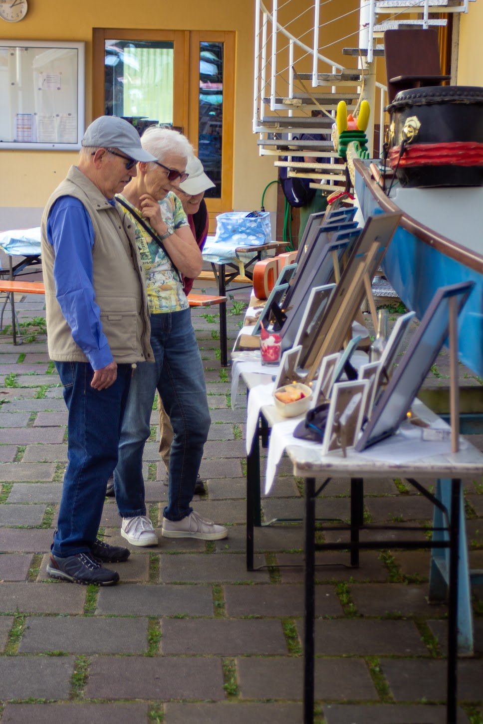 Drei ältere Leute betrachten einen Stand mit Bildern und Dekorationen bei einem Flohmarkt im Freien.