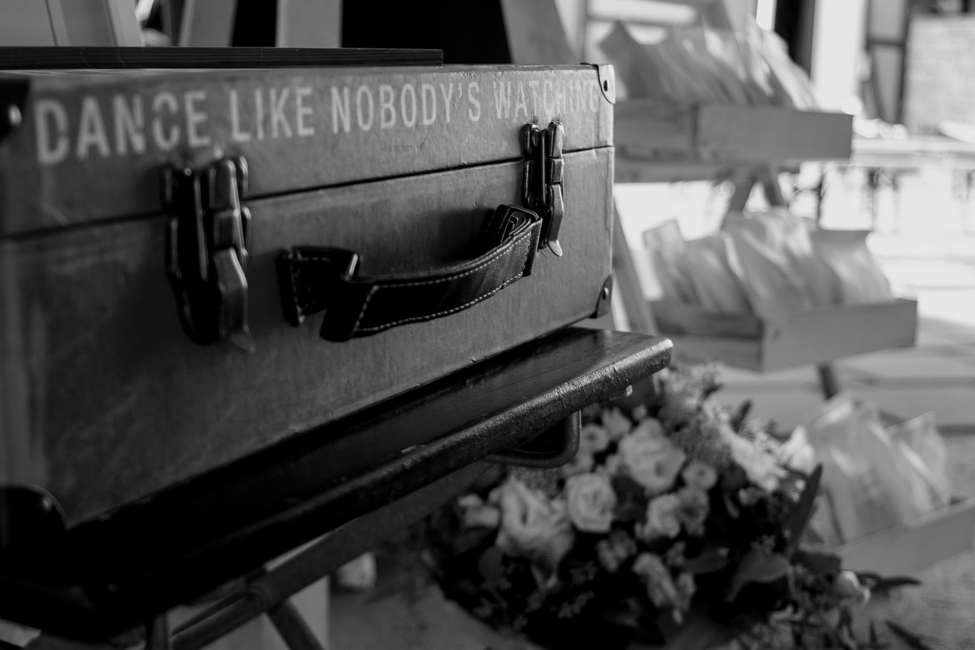 Schwarzes Eisenkoffer mit weißen Aufdruck, auf einer Holzbank, im Hintergrund Blumen und Verkaufsstände sichtbar.