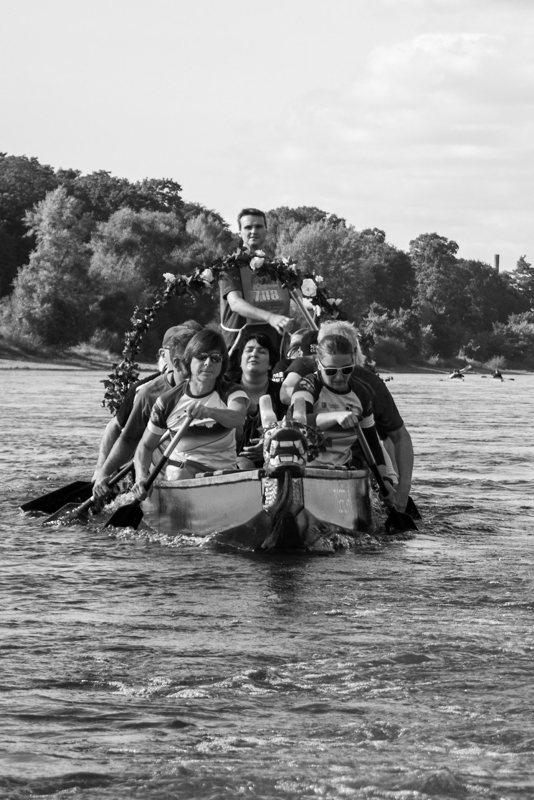 Eine Gruppe von Menschen paddelt in einem Drachenboot auf einem Fluss, mit einem Mann am Steuer, im Hintergrund Bäume und andere Boote.