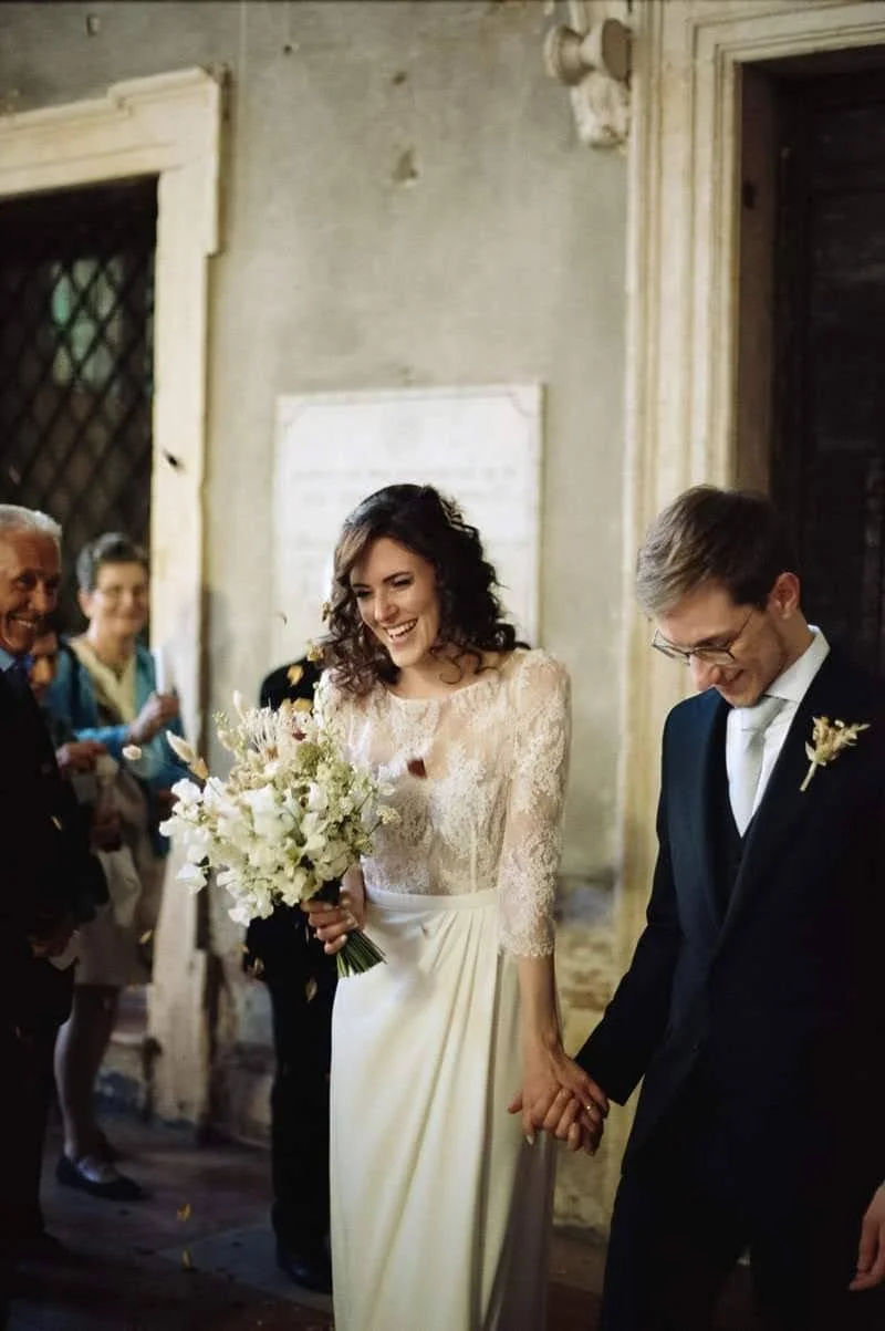 Ein glückliches Paar bei einer Hochzeit, die Braut trägt ein weißes Kleid und hält einen Blumenstrauß, der Bräutigam trägt einen schwarzen Anzug, sie halten sich an den Händen und lächeln, im Hintergrund feiern Gäste die Zeremonie.