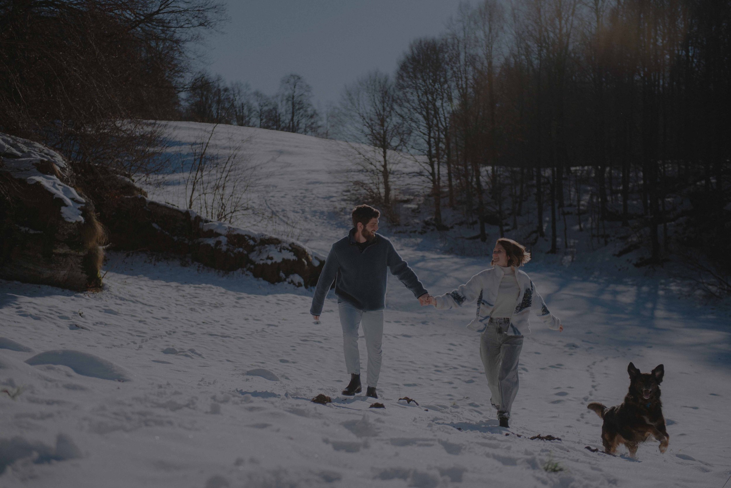 Winterliches Paarshooting in den Bergen mit Hund, Hand in Hand im Schnee während der goldenen Stunde. Winter couple photoshoot in the mountains with their dog, walking hand in hand through the snow during golden hour.