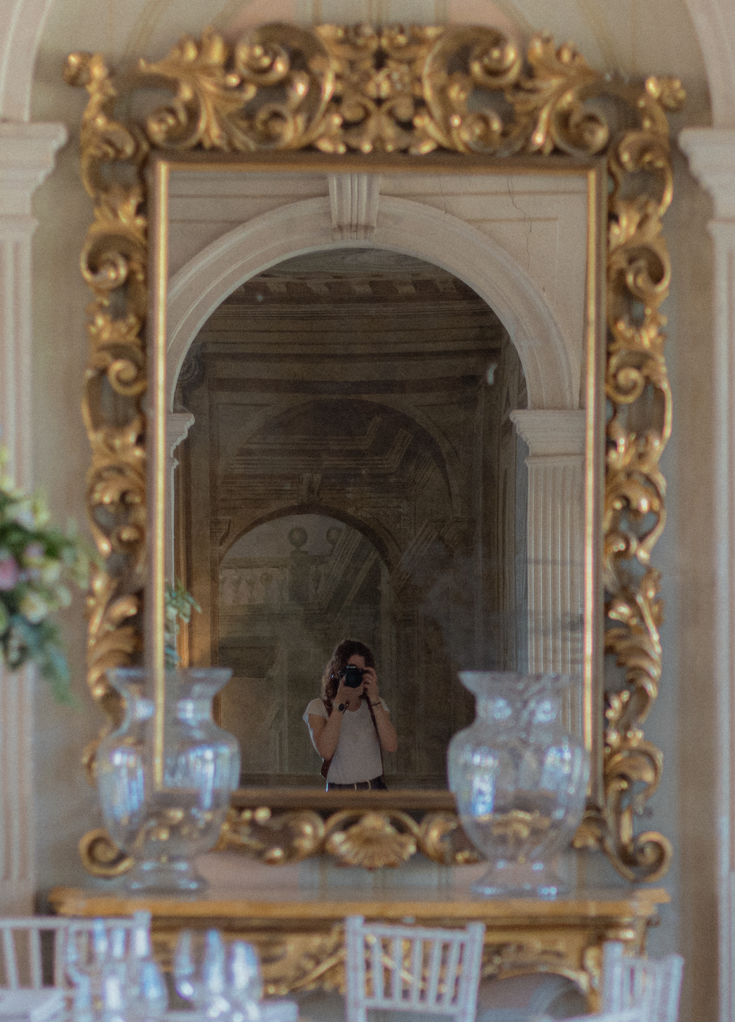 Ein Spiegel mit einem goldfarbenen, kunstvollen Rahmen, der eine Person beim Fotografieren zeigt, im Hintergrund eine prunkvolle Decke und Säulen in einem eleganten Raum.