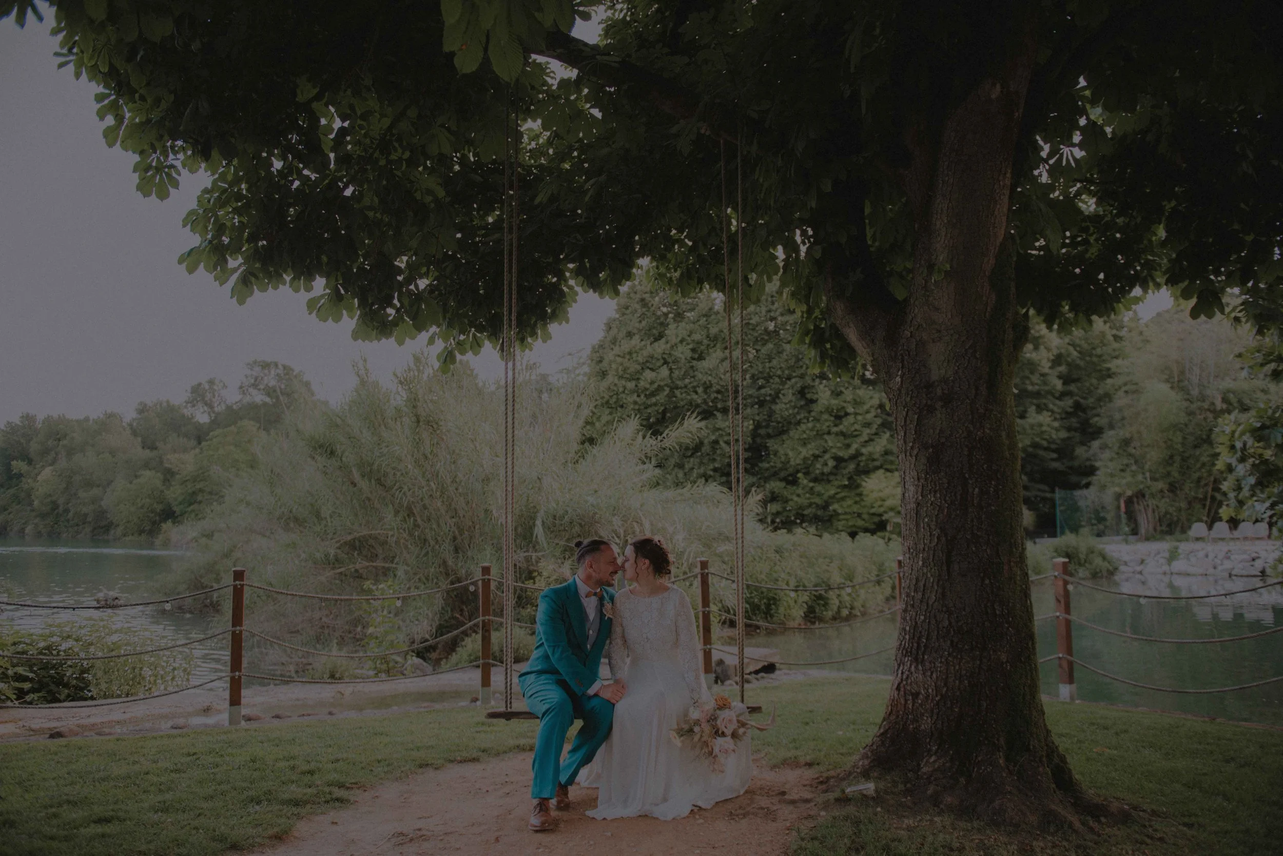 Romantische Sommerhochzeit in Verona, Italien, mit Braut im Spitzenkleid und Bräutigam im petrolfarbenen Anzug auf einer Schaukel am See. Romantic summer wedding in Verona, Italy, with bride in a lace gown and groom in a teal suit sitting on a swing.