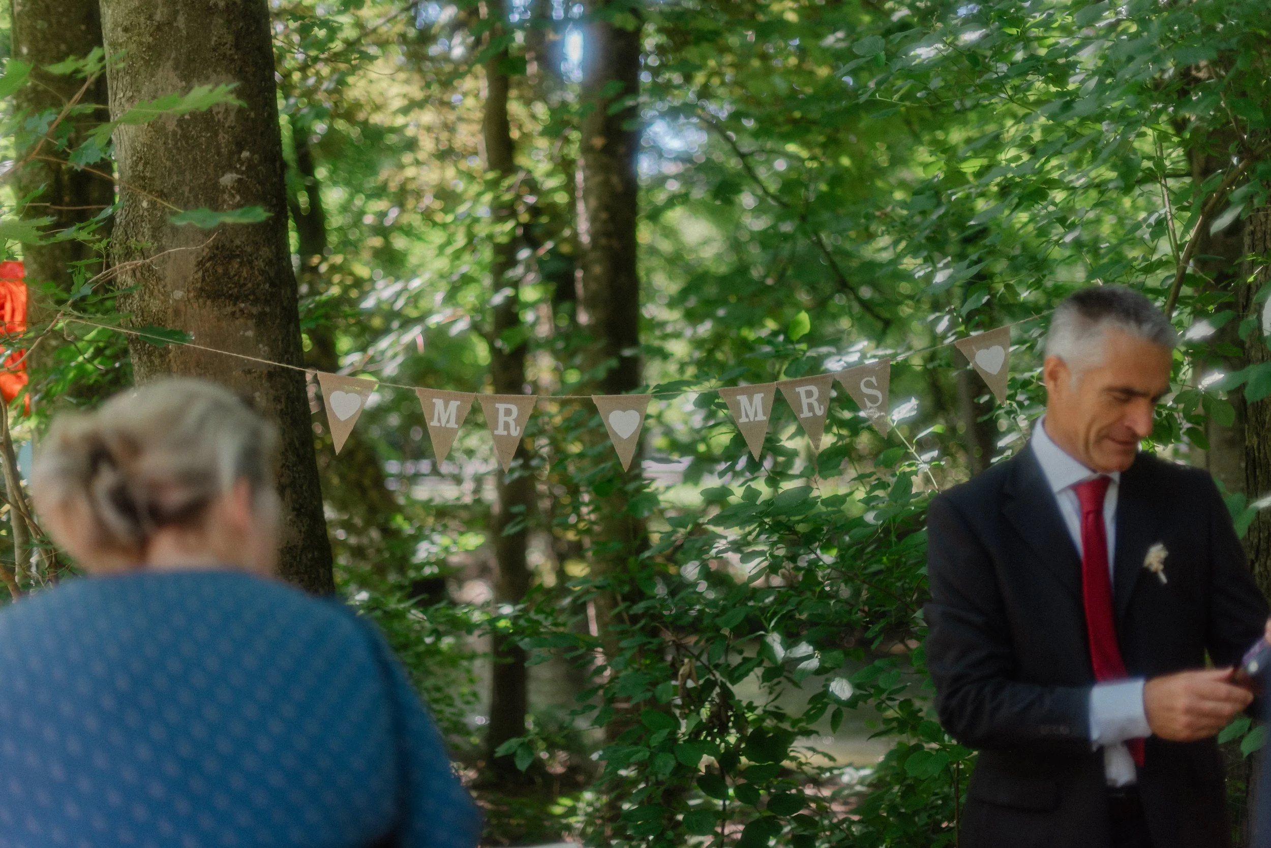 Ein Mann und eine Frau stehen im Wald, hinter ihnen eine Schnur mit einem Banner, auf dem ‚MARRIAGE’ steht, und Herzen. Es ist wahrscheinlich eine Hochzeit im Freien.