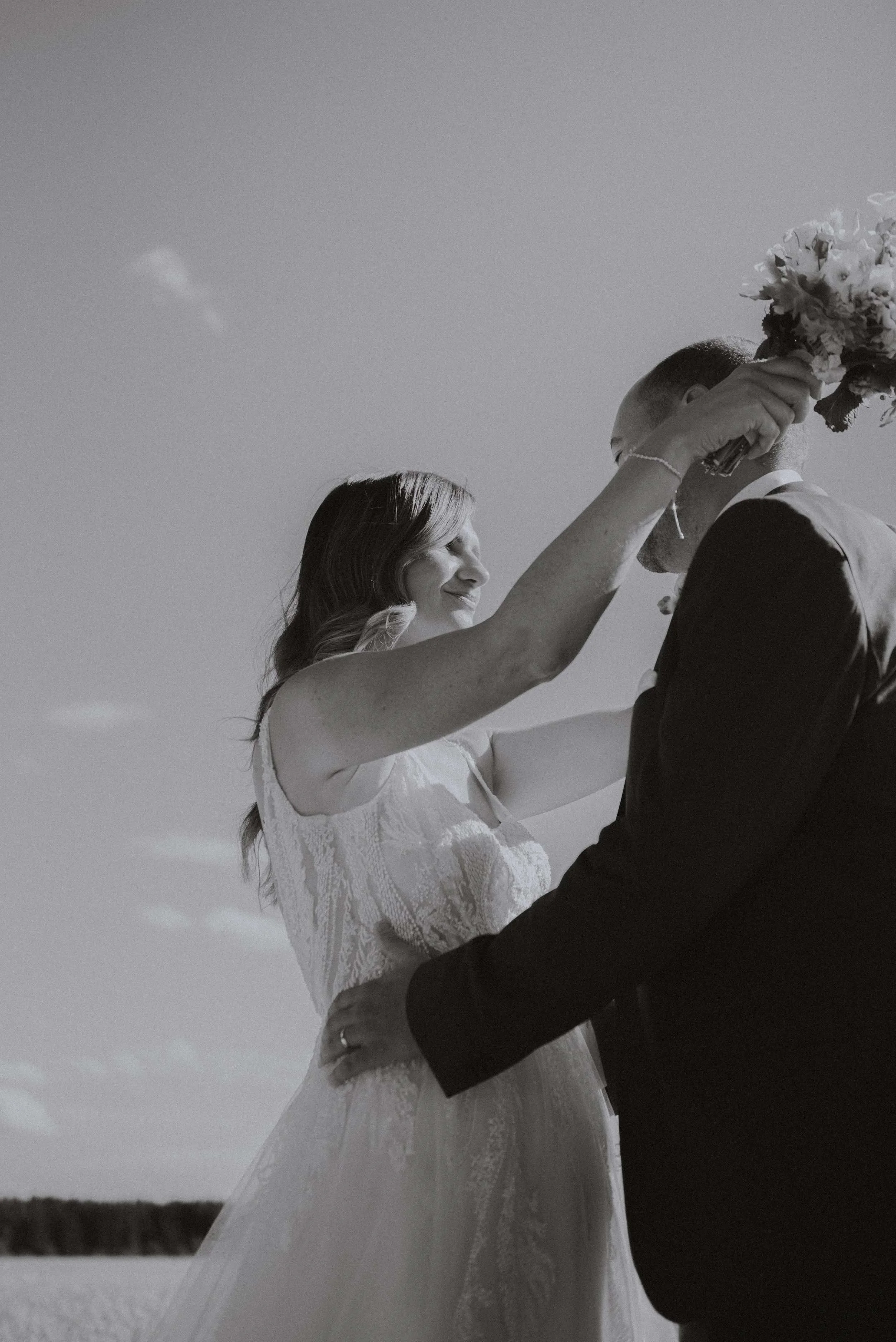 Braut und Bräutigam bei ihrer Hochzeit im Freien, in Schwarz-Weiß, der Braut hält einen Blumenstrauß in der Hand. Es ist ein klarer Himmel im Hintergrund.
