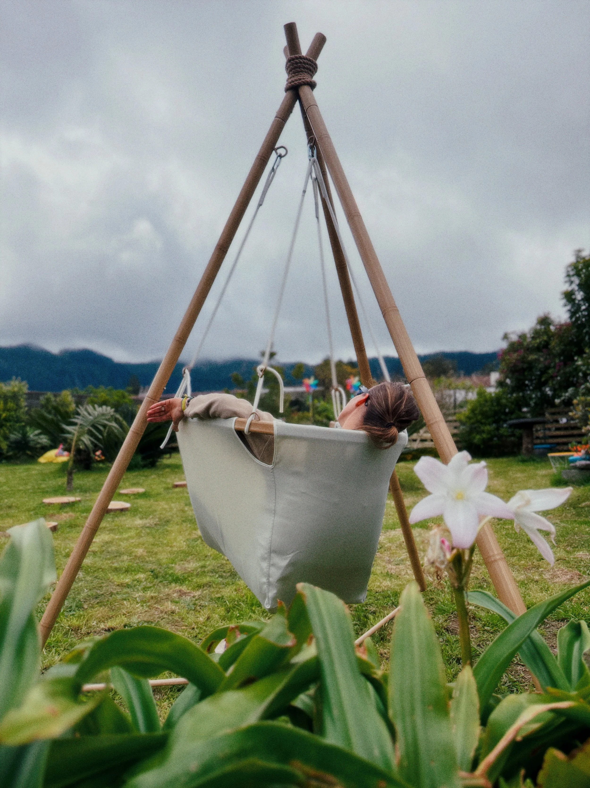 A woman reclining in a swing seat outdoors in a garden, surrounded by green plants and flowers, with mountains and cloudy sky in the background.