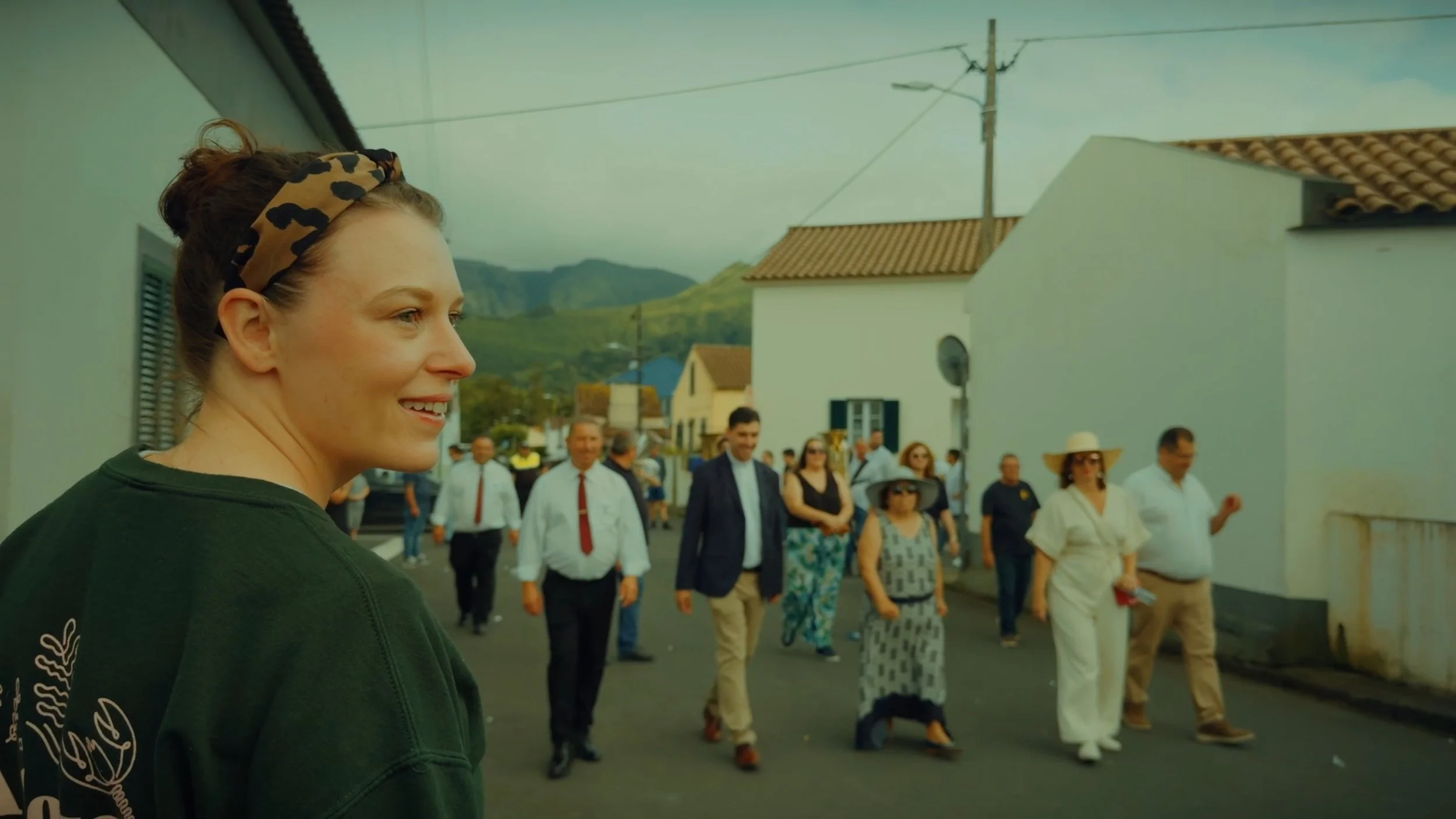 A woman with a leopard-print headband smiling in profile, with a group of people walking behind her on a street in a town with mountains in the background.