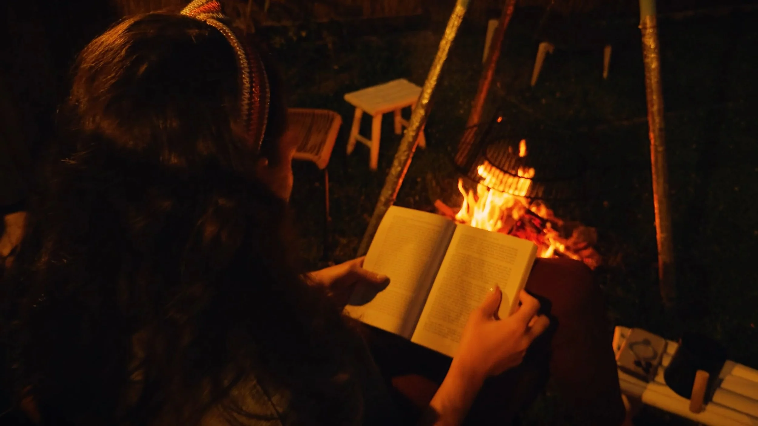 Person reading a book near a campfire at night with outdoor chairs and a small table in the background.