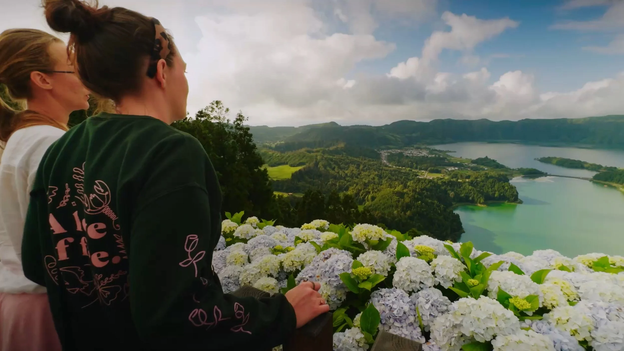 Three women sitting outdoors overlooking a scenic landscape with a river, hills, and partly cloudy sky, surrounded by white hydrangea flowers.