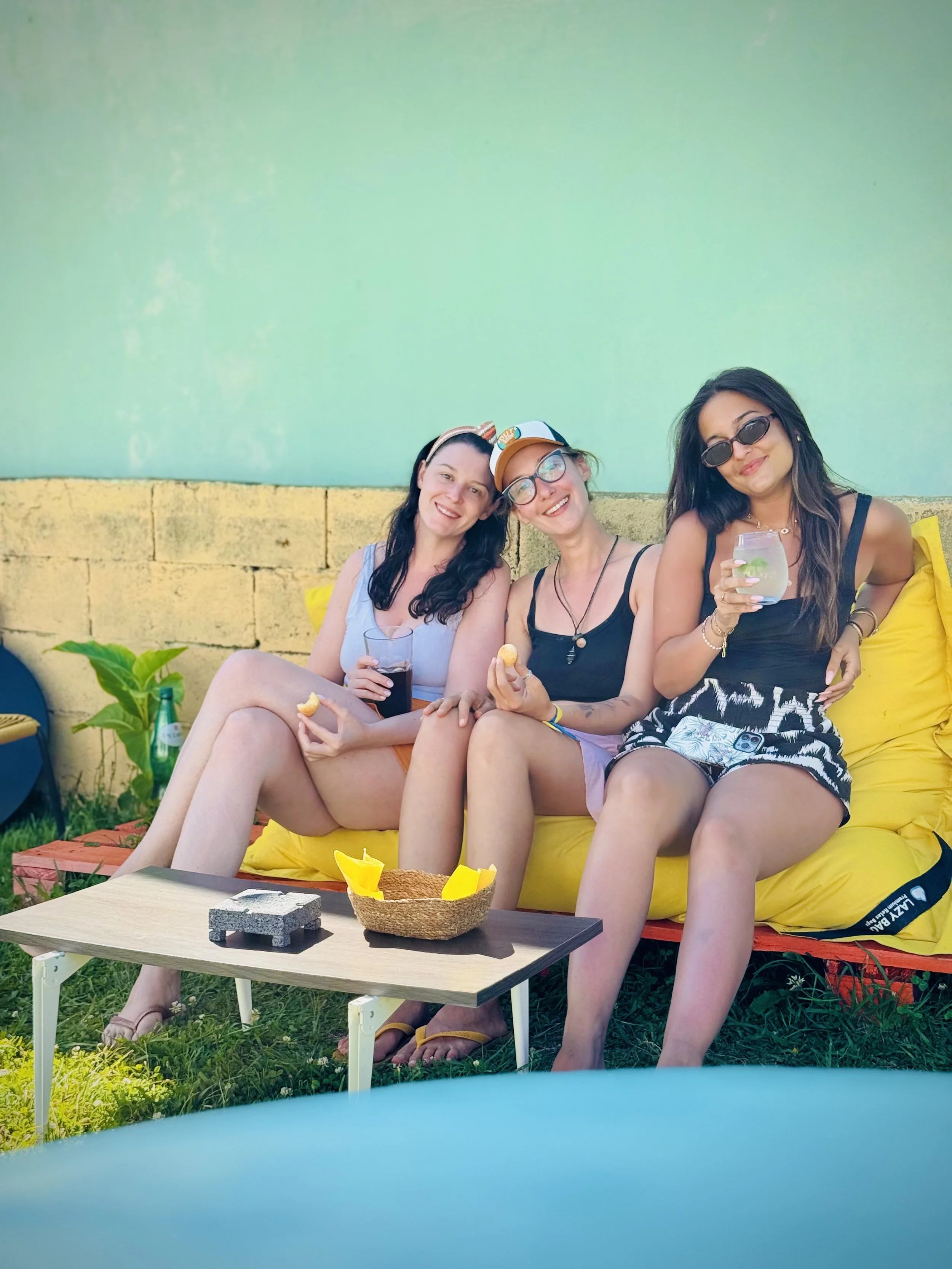 Three women sitting outdoors on a yellow couch, enjoying drinks and snacks, with a brick wall and green plants behind them.