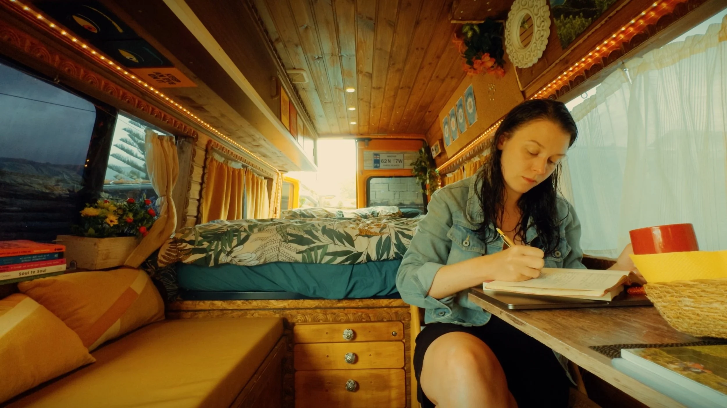 A young woman with wet hair sitting at a table inside a cozy camper van, writing in a notebook, with books, plants, and colorful decor around her.
