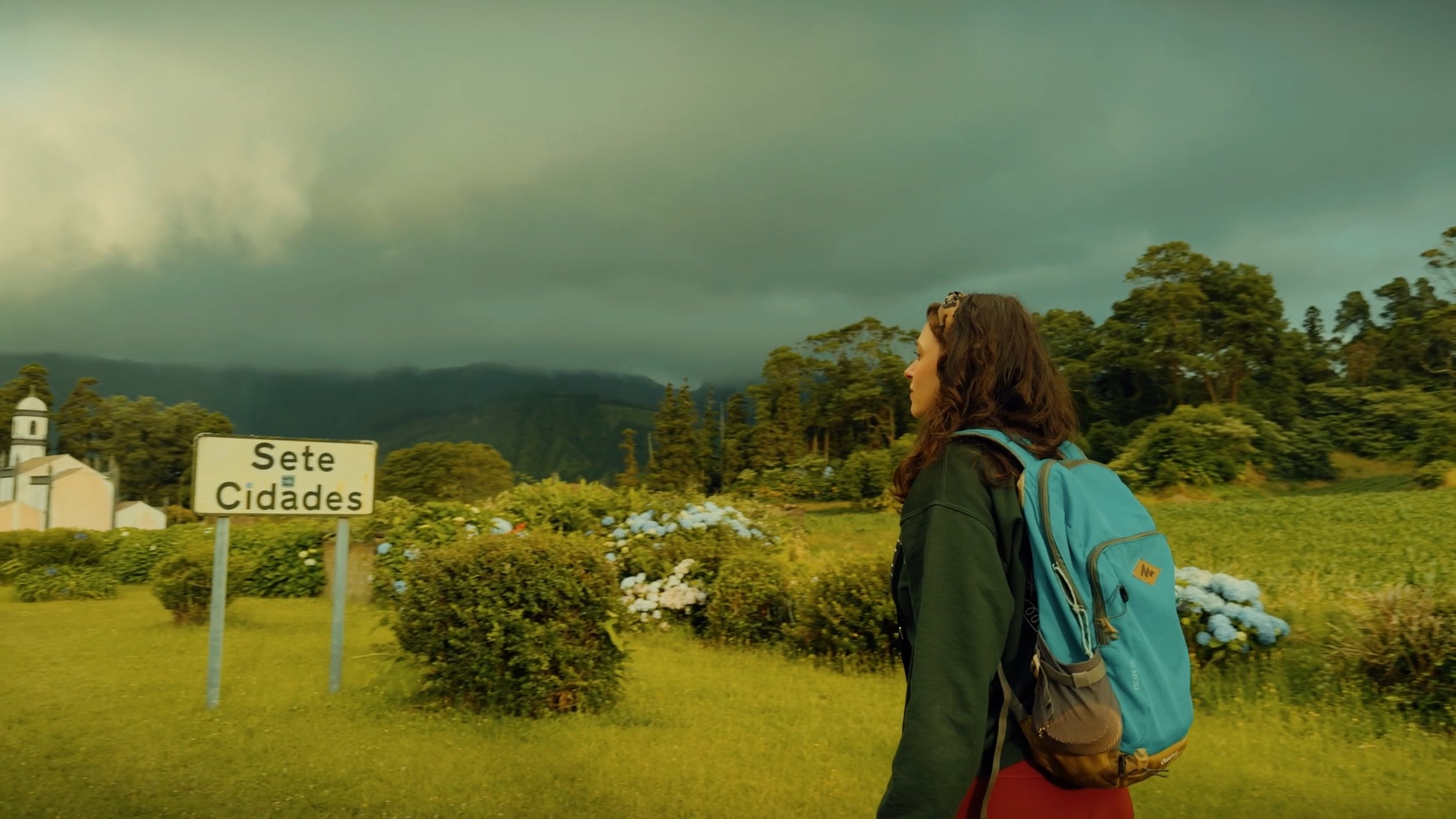 A woman with long curly hair wearing a black jacket and a large blue backpack standing in front of a sign that reads "Sete Cidades," with trees and dark clouds in the background.