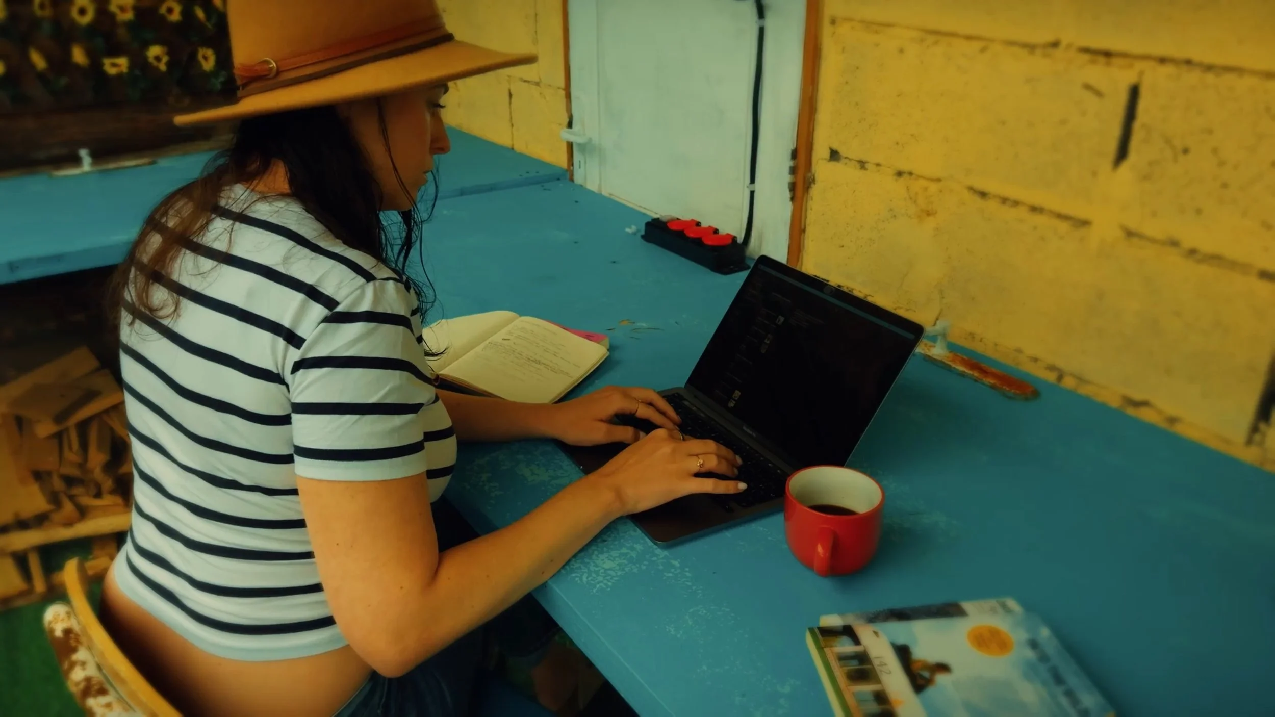 A woman wearing a striped shirt and a wide-brimmed hat sitting at a blue table, working on a laptop with an open notebook beside her. There is a red mug with dark beverage on the table. The setting appears to be indoors with yellow brick walls.