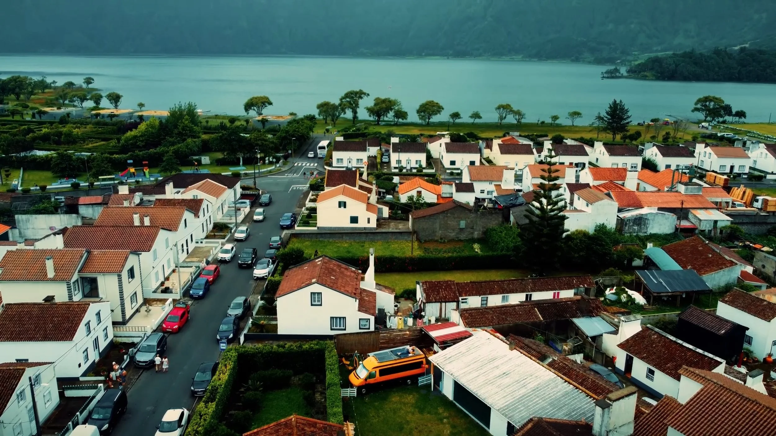 Aerial view of a residential neighborhood with white houses featuring red-tiled roofs, parked cars along streets, a lake in the background, and green hills and trees surrounding the area.
