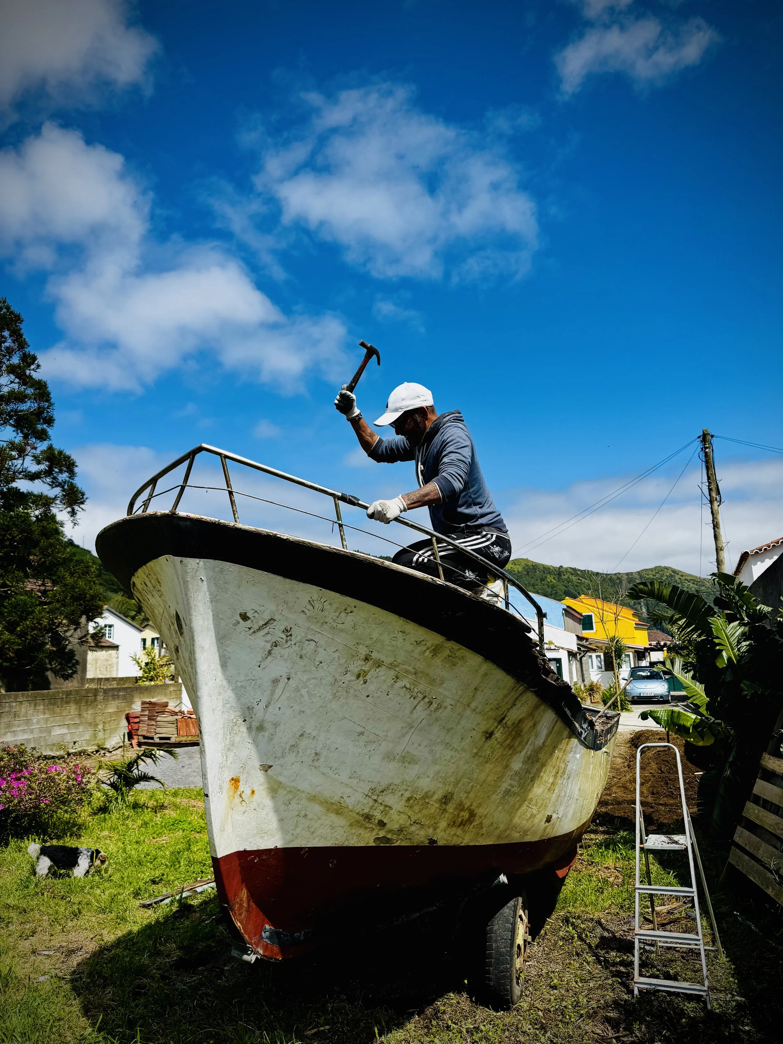 A man wearing a white cap and gray hoodie hammering on a boat in a yard with houses, trees, and a blue sky with clouds in the background.