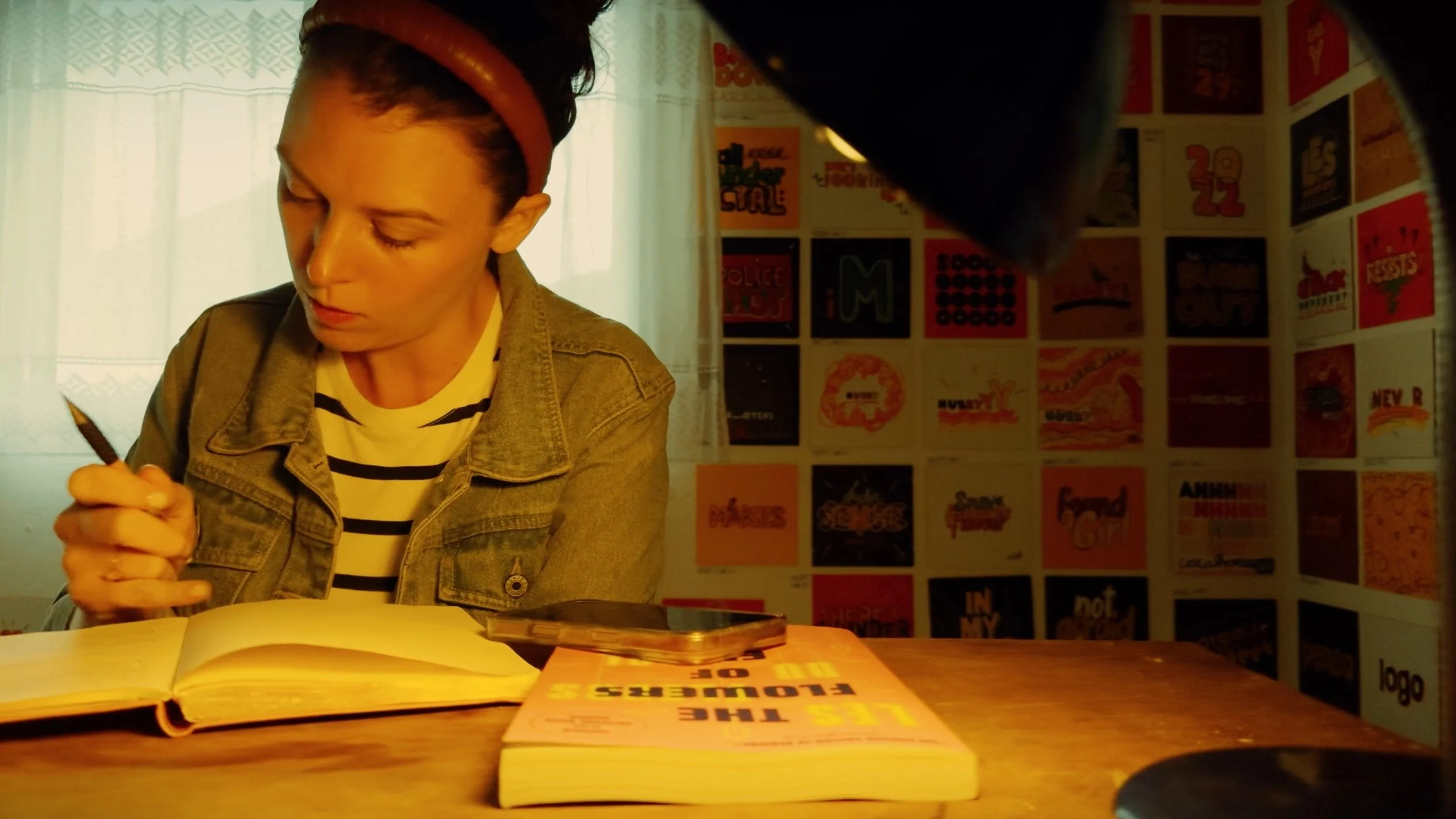 A woman with short dark hair wearing a striped shirt and jean jacket sitting at a table, reading a book with colorful posters on the wall in the background.