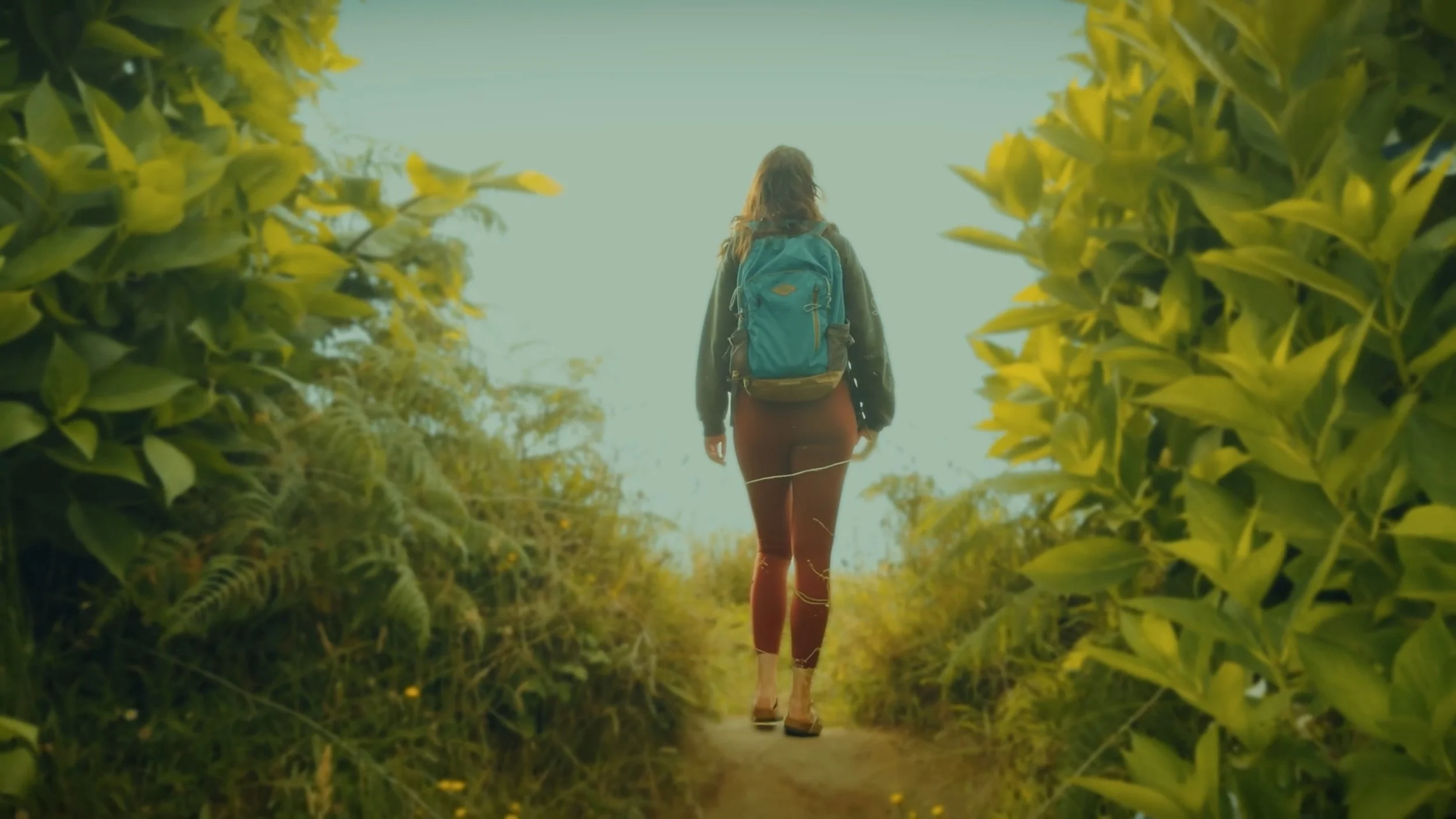 A person walking through a lush green pathway with foliage on both sides, carrying a backpack and dressed casually.