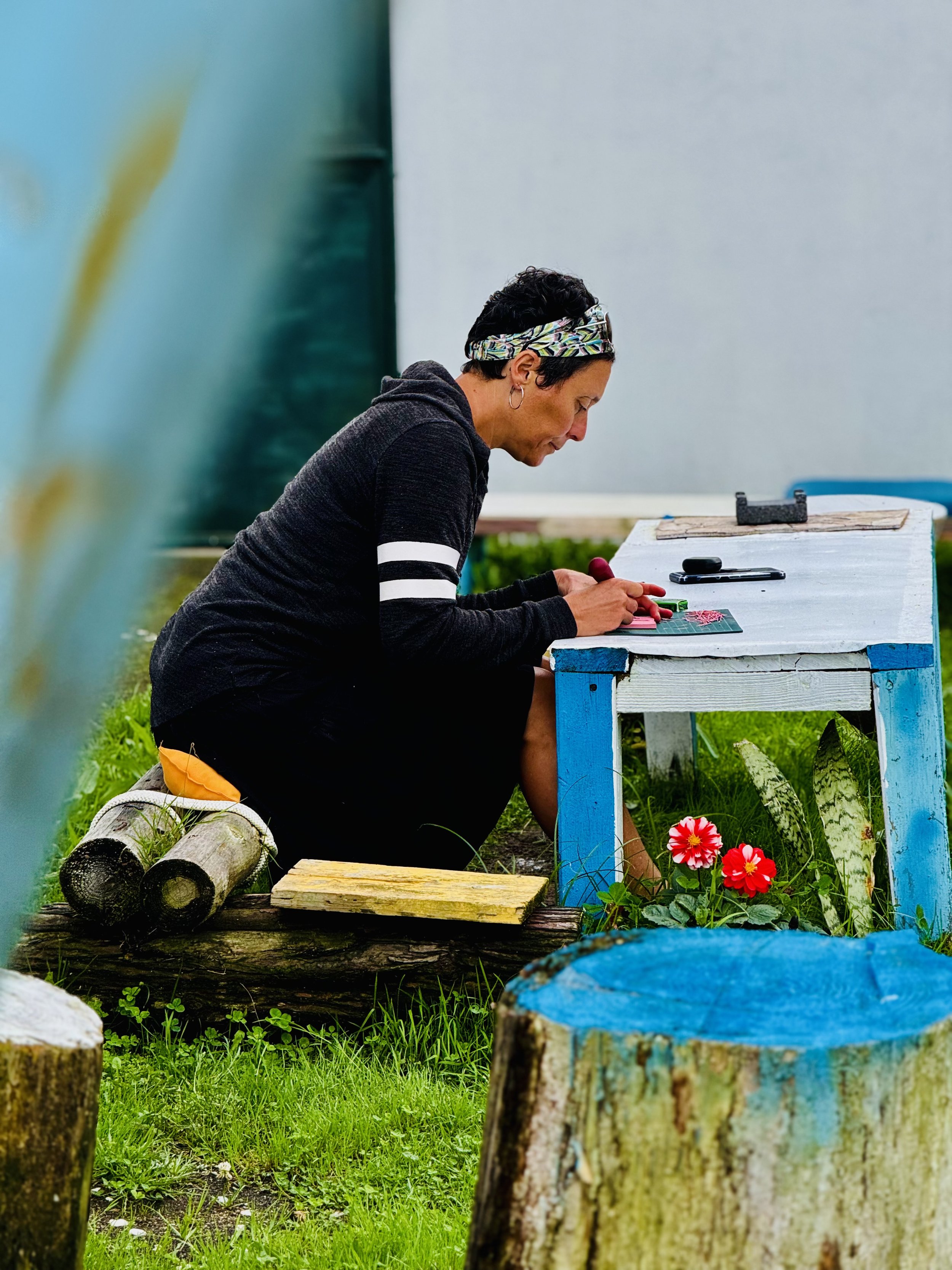 Woman with short black hair, wearing a black hoodie with white stripes, working on a craft project at an outdoor workbench with plants and colorful tree stumps nearby.