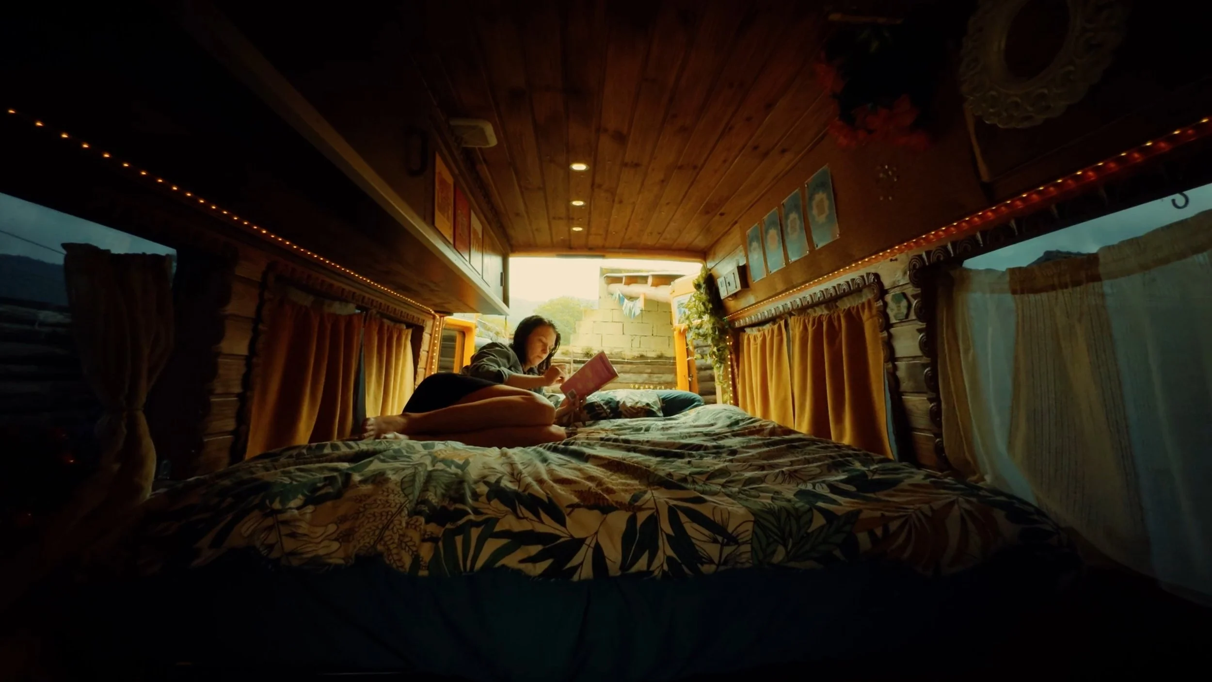 Person lying on a bed inside a cozy wooden camper, reading a book during the daytime with sunlight coming through the window.