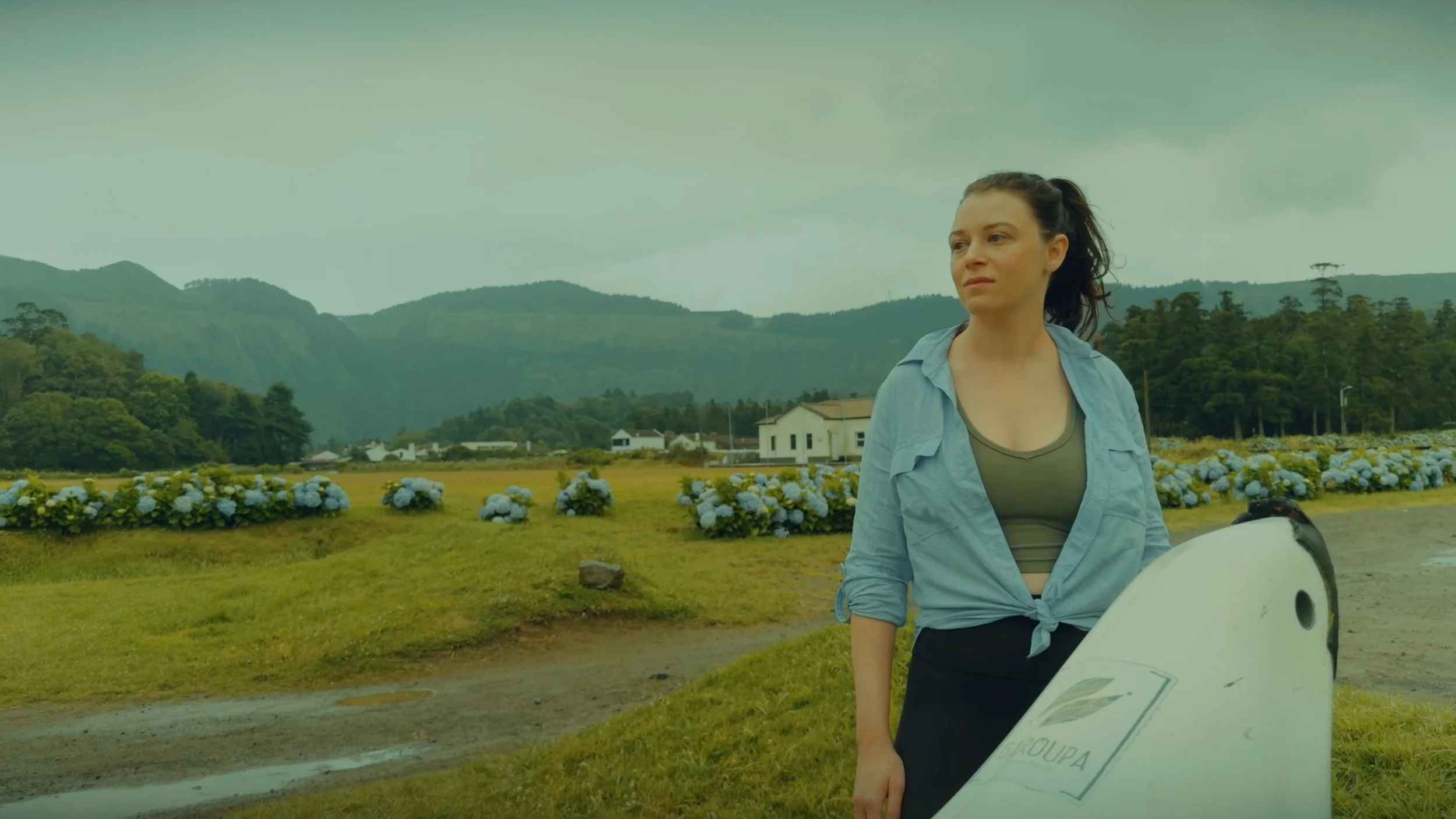 A woman stands outdoors near hydrangea flowers, holding a surfboard, with mountains and a cloudy sky in the background.
