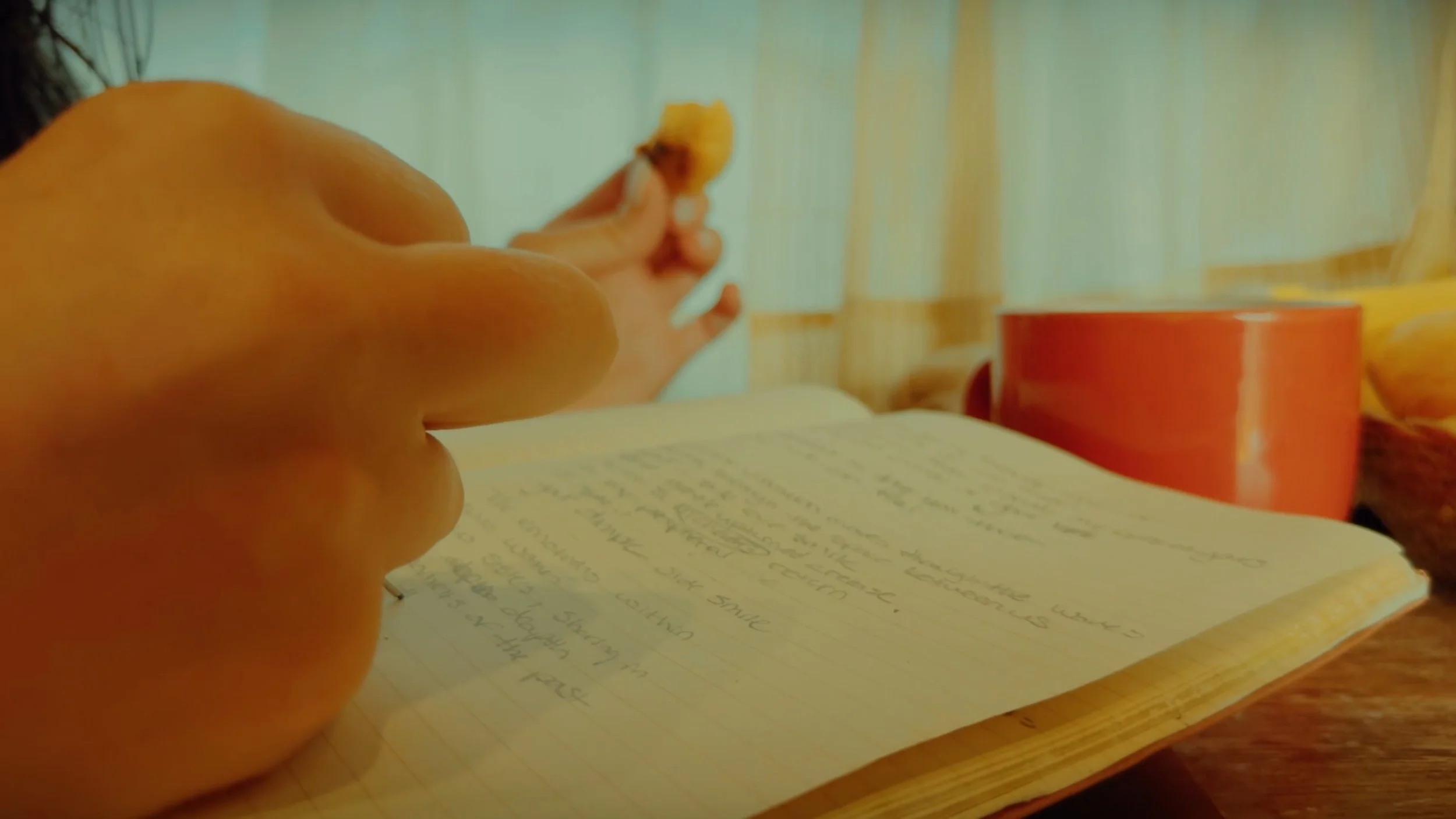 Close-up of a person's hand holding a piece of bread or a snack above an open handwritten journal or notebook on a wooden table. In the background, there is a red mug and some yellow objects, possibly food or packaging, near a window with blinds.