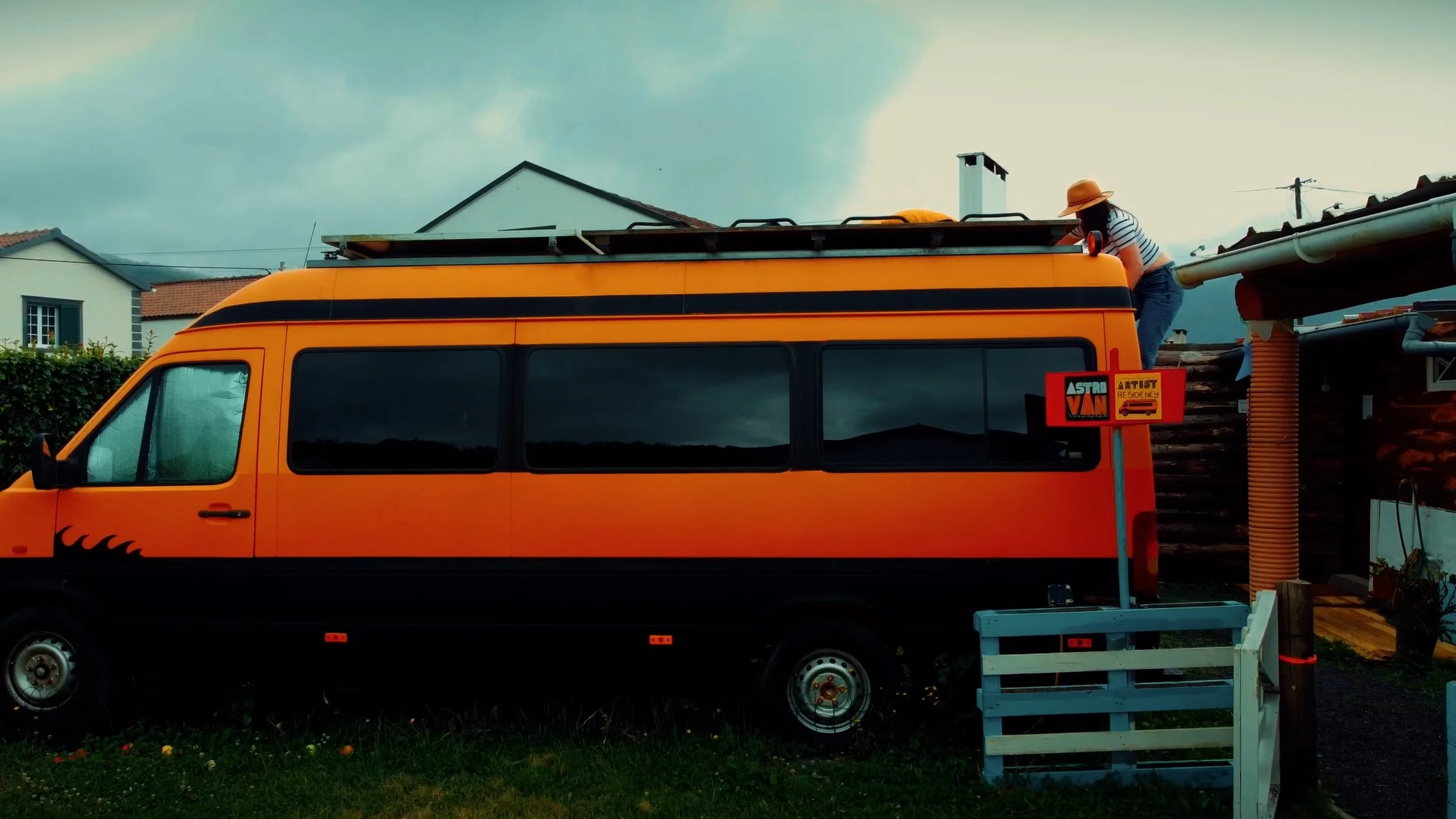 A bright orange van parked in a yard with a woman in a striped shirt and orange hat standing on its roof, working on the roof or installing something. There is a sign on the van that reads 'Astro Van Artist Residency' and a small white fence in front