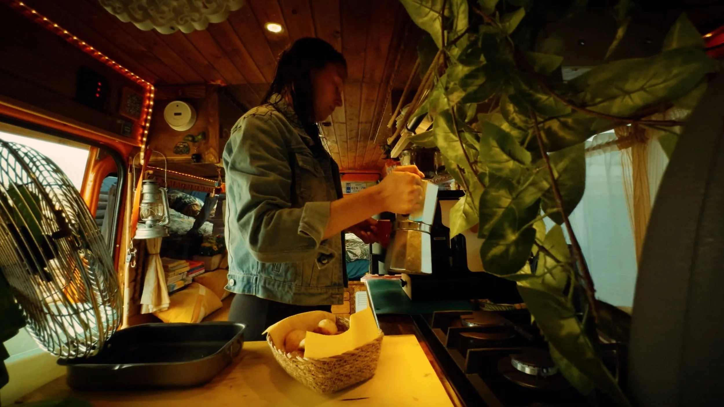 A woman with dark, wet hair wearing a denim jacket is writing on a notepad inside a cozy, wood-paneled room decorated with plants, books, and warm lighting.