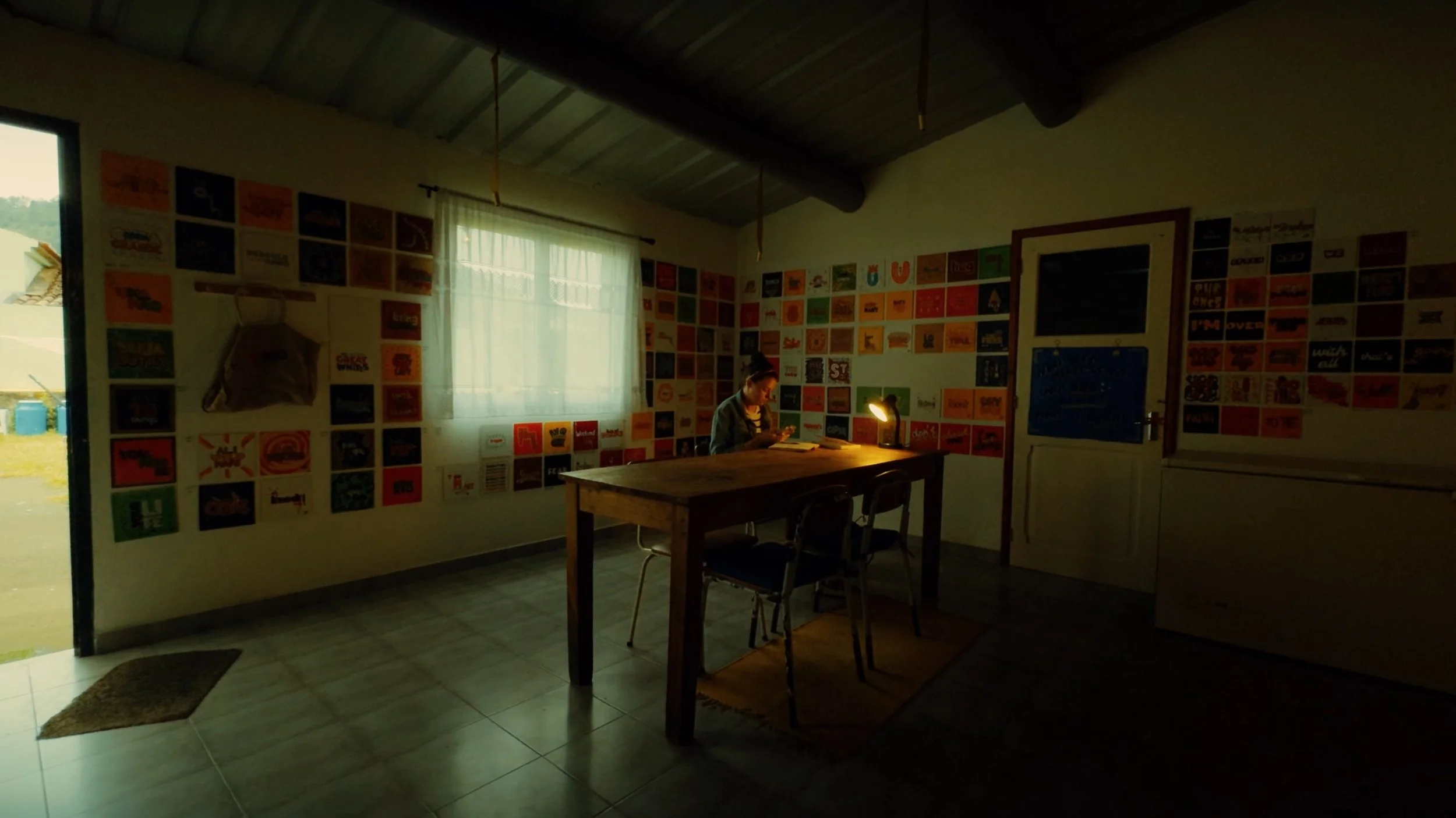 A woman is sitting at a wooden table inside a room with walls decorated with numerous framed pictures and artwork. She appears to be reading or looking at something in her hands. There is a window with sheer curtains behind her and a table lamp provi