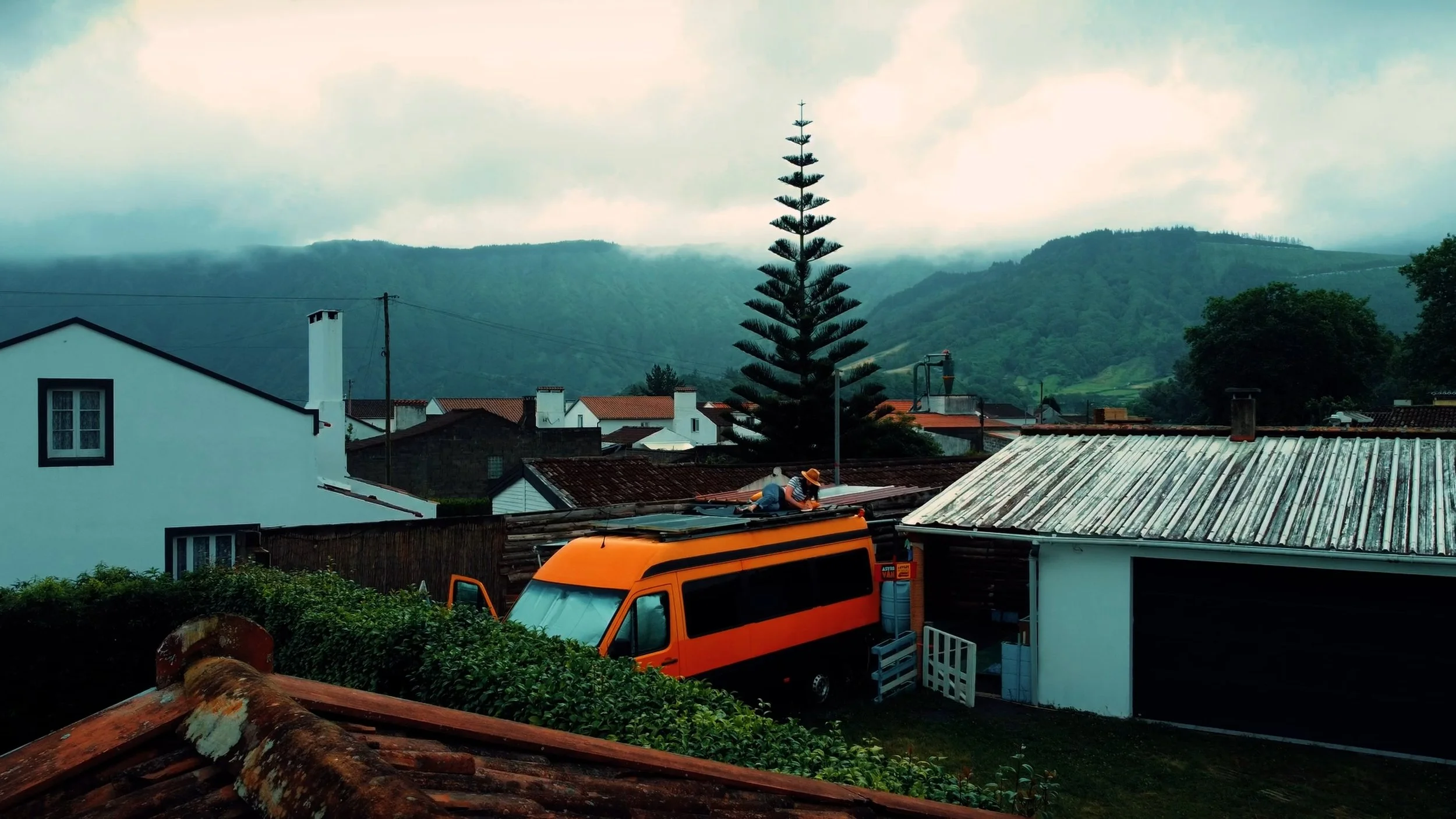 A rural neighborhood with white houses, a large pine tree, mountains in the background, and a worker on the roof of a house in the foreground.