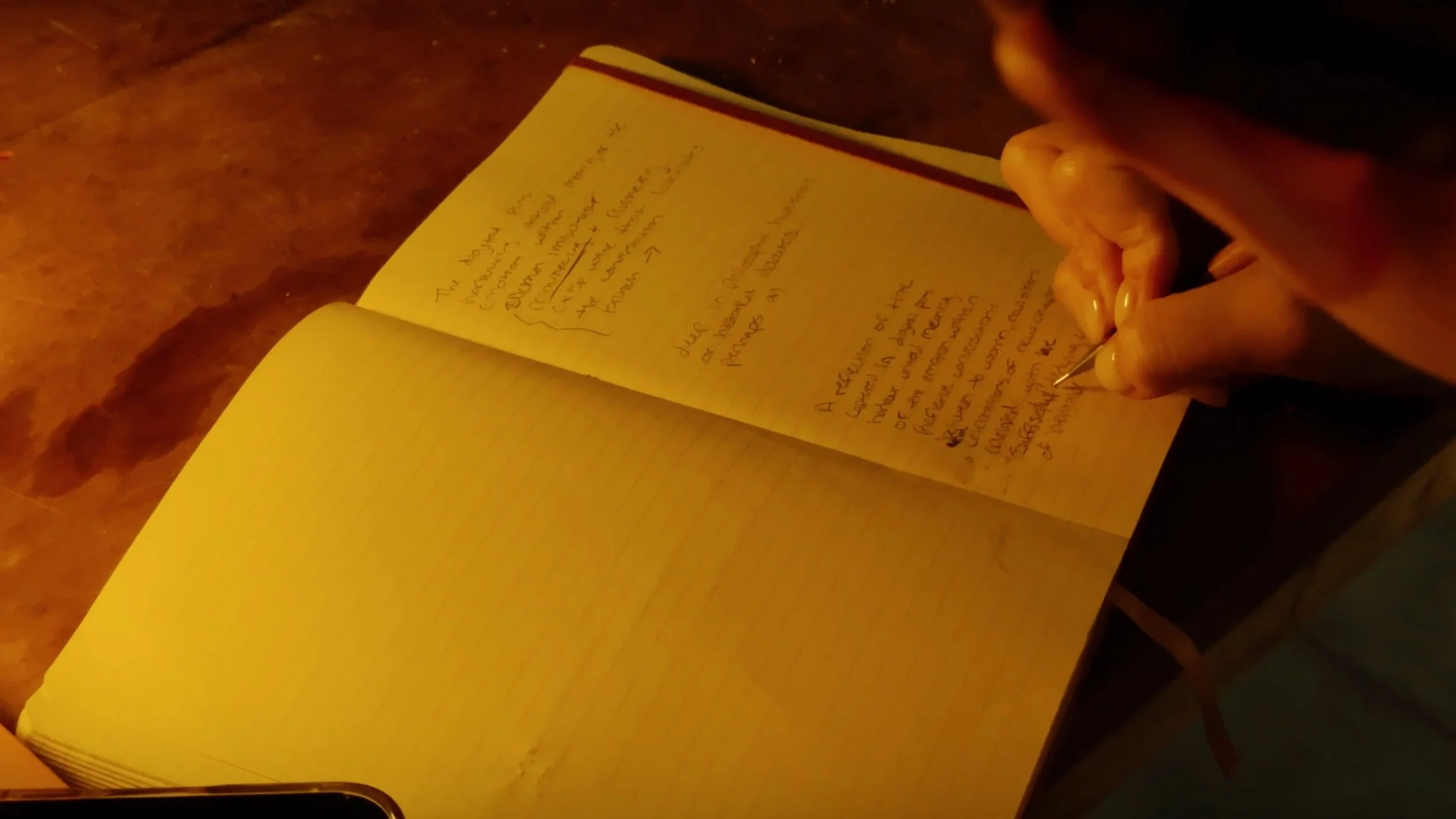A person writing notes in a lined notebook, with handwritten text visible on the pages, under warm lighting.