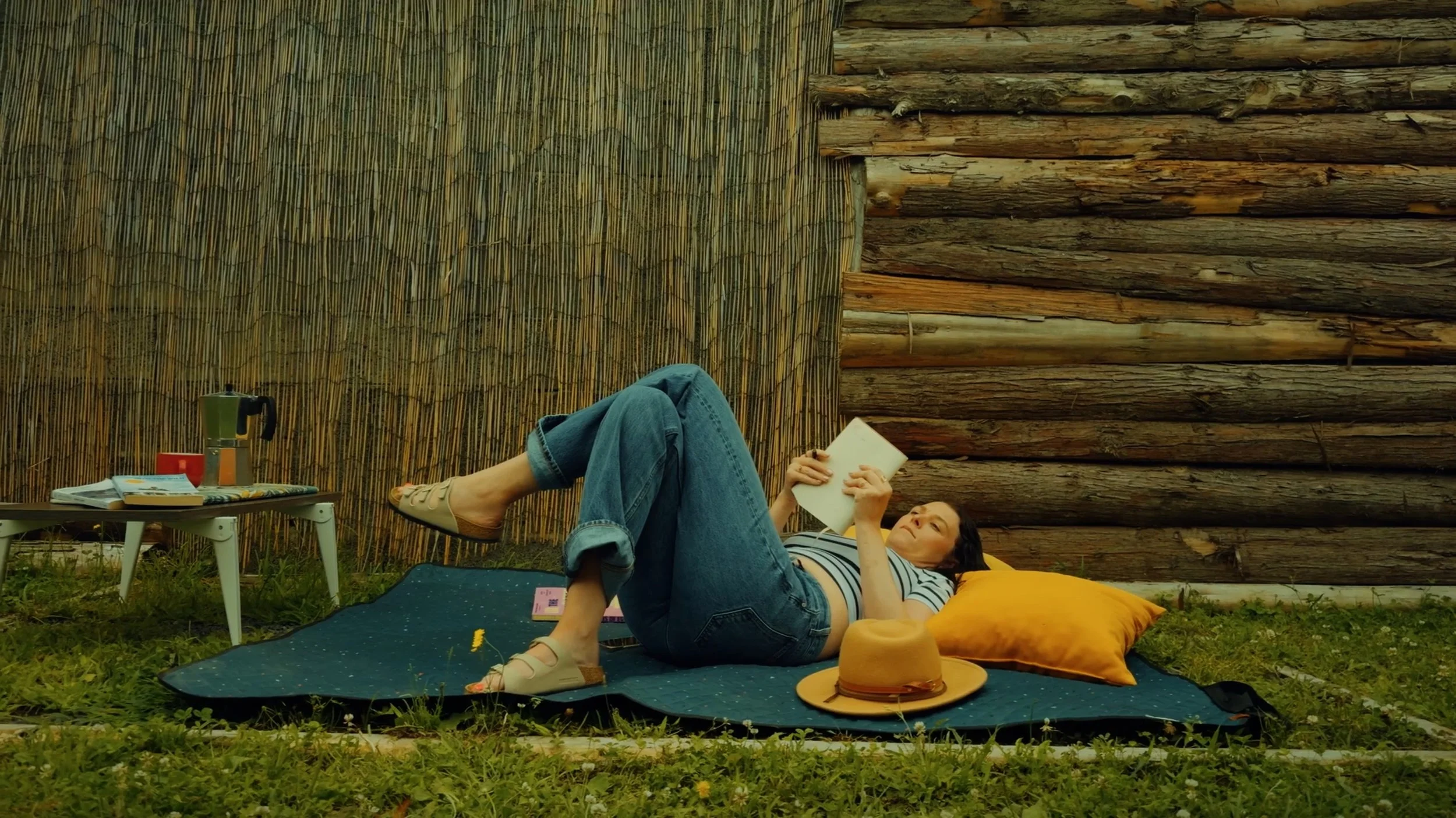 A woman lying on a black outdoor rug reading a book with her legs resting on a yellow pillow outdoors. She is wearing a striped shirt, jeans, and sandals. There is a small white table with a coffee pot, mug, and magazines nearby, and a straw hat on t