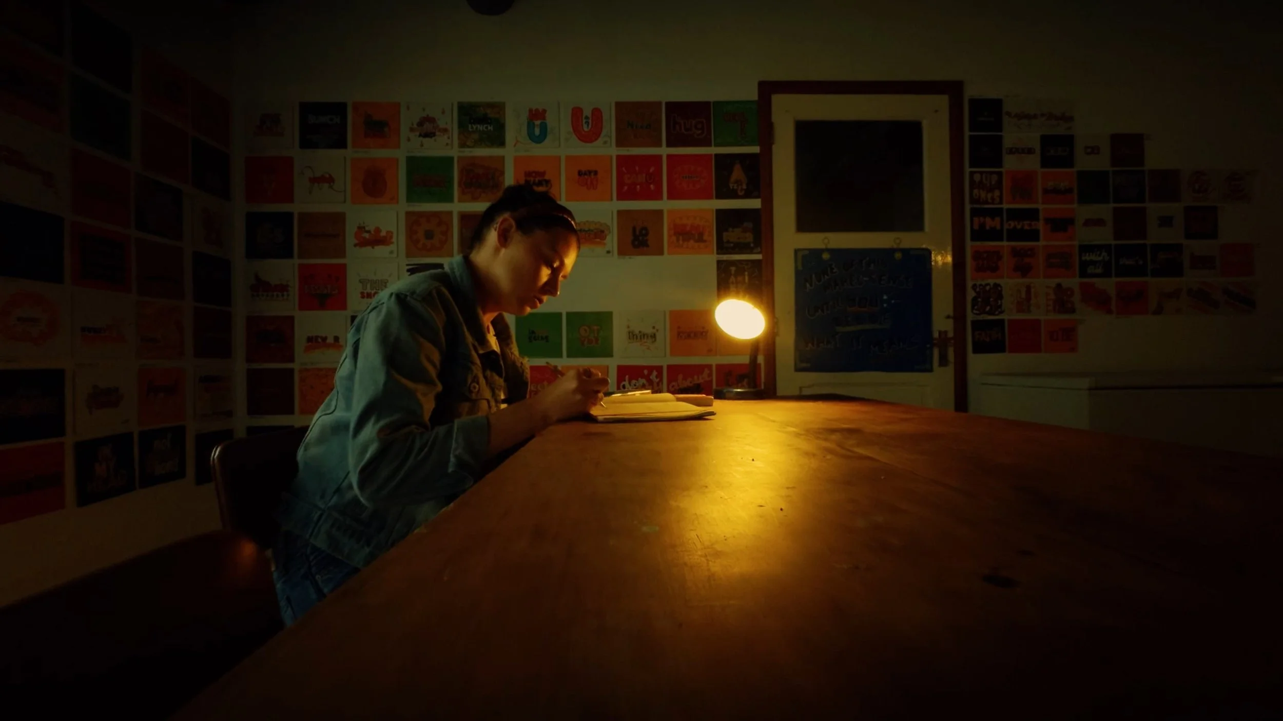 Person sitting at a wooden table reading a book, illuminated by a small yellow lamp, in a dark room with colorful artwork on the walls.