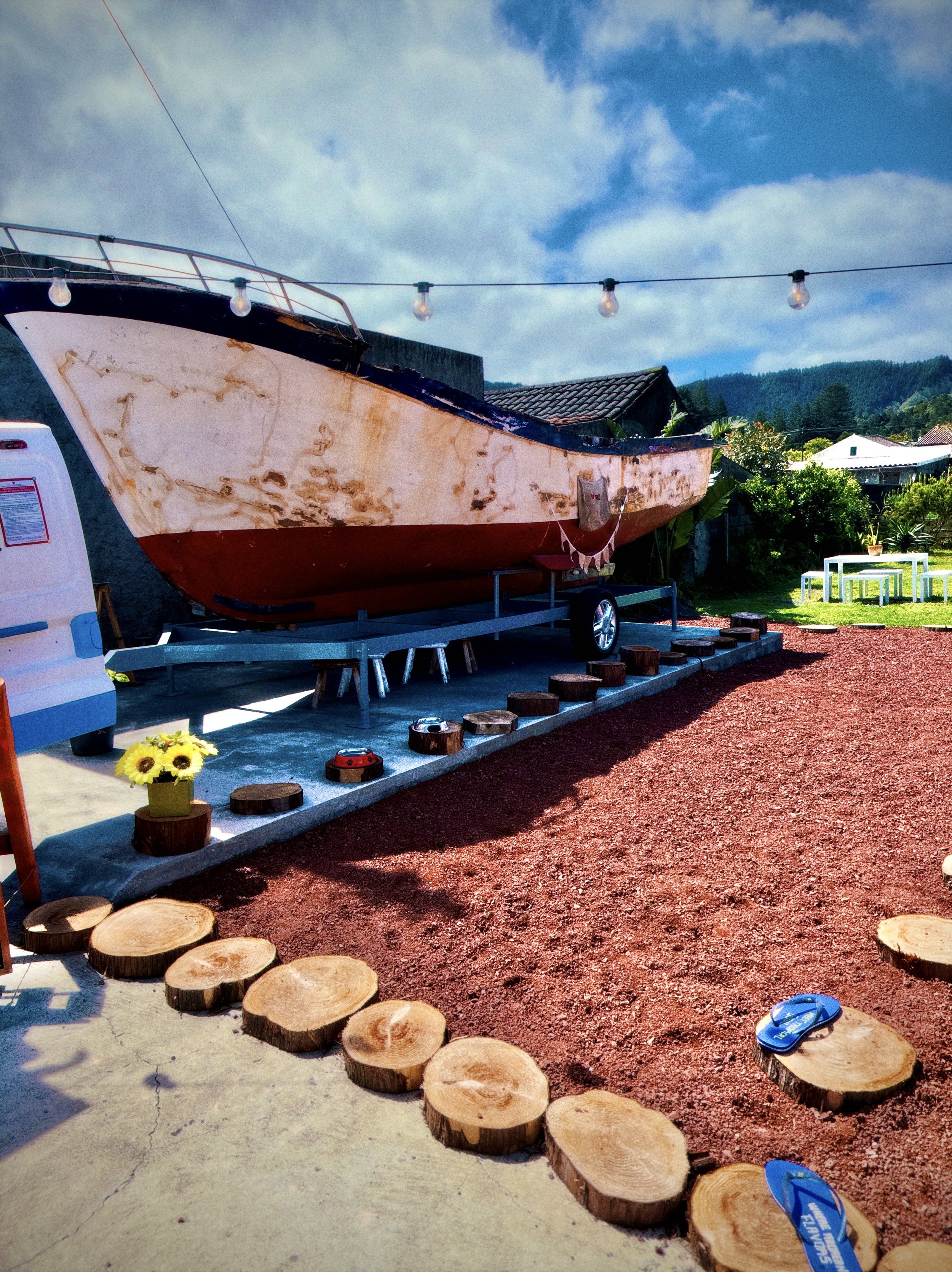 An outdoor scene featuring a boat on a trailer, with a string of lights overhead, a small table with plants, and groups of slices of wood, along with a pair of blue flip-flops.
