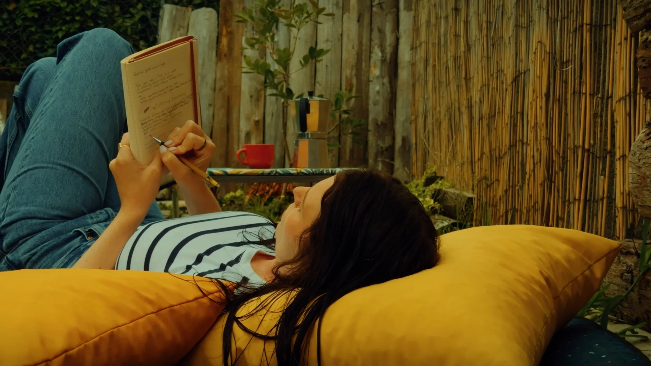 Woman lying on a yellow cushion reading a notebook outdoors, with another person sitting nearby.