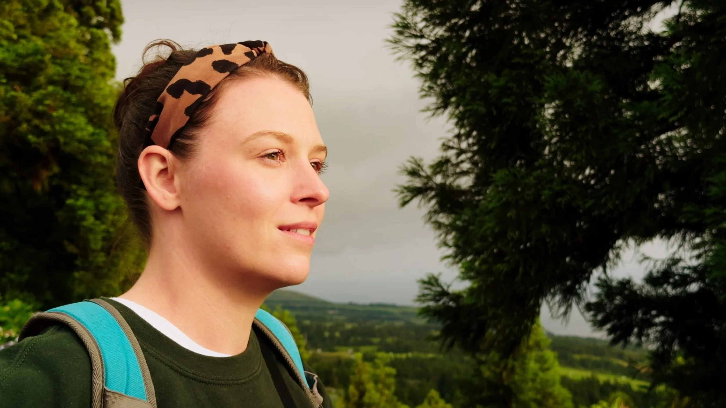 A young woman with a leopard print headband and a backpack, standing outdoors near trees and hills.