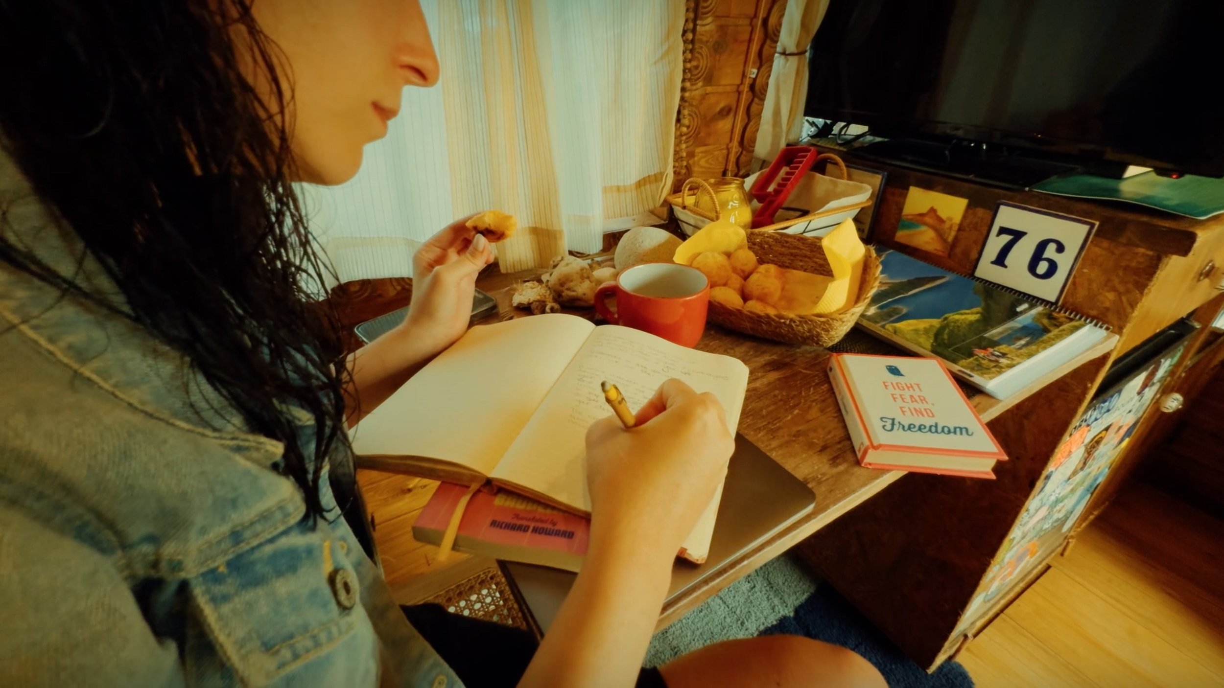 A woman with curly dark hair reading a notebook at a wooden table filled with snacks, a cup, books, and pictures, in a cozy room with wooden walls and a TV in the background.
