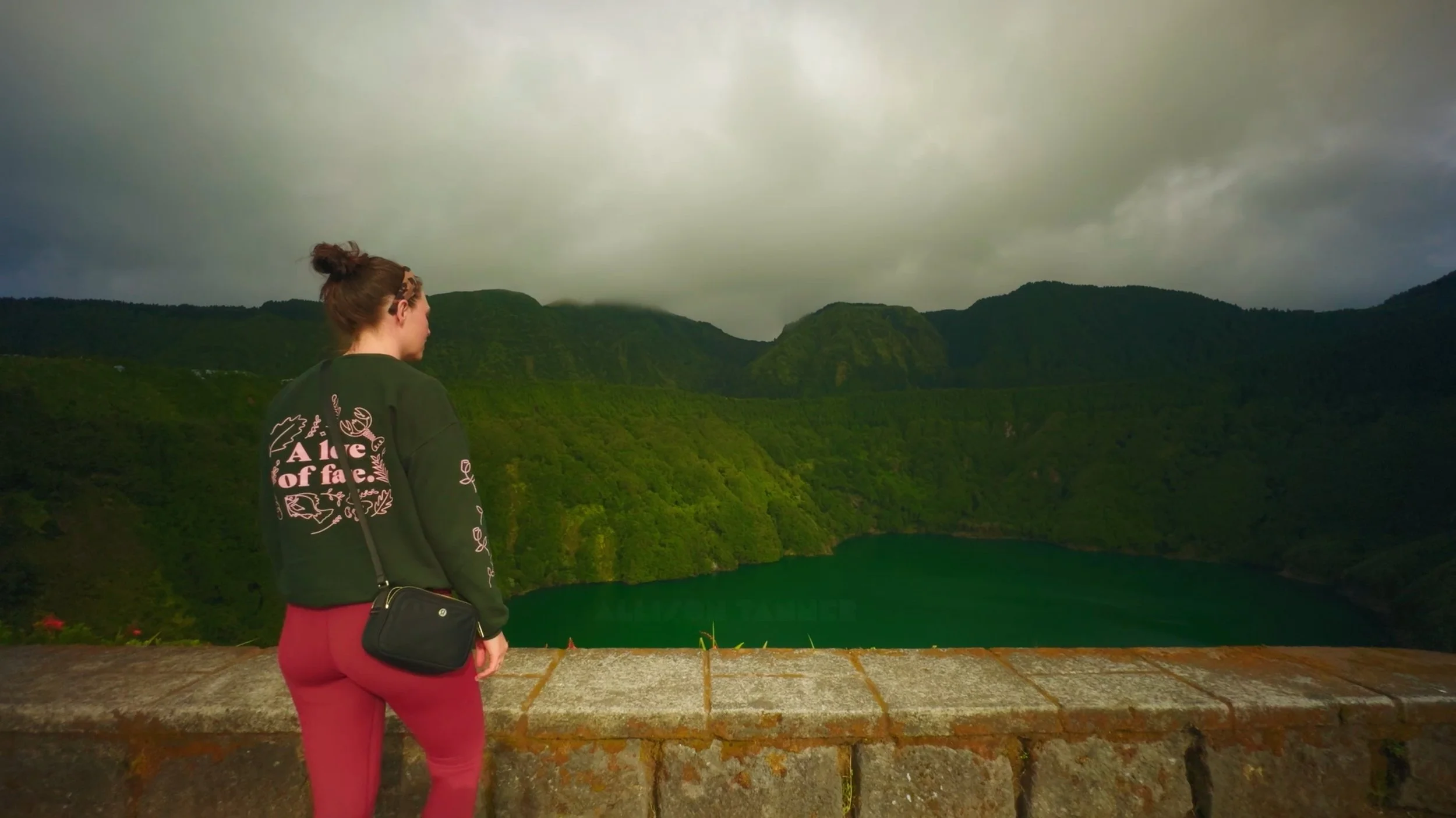 A woman standing at a lookout overlooking a lush green landscape with rolling hills and a body of water, cloudy sky overhead.
