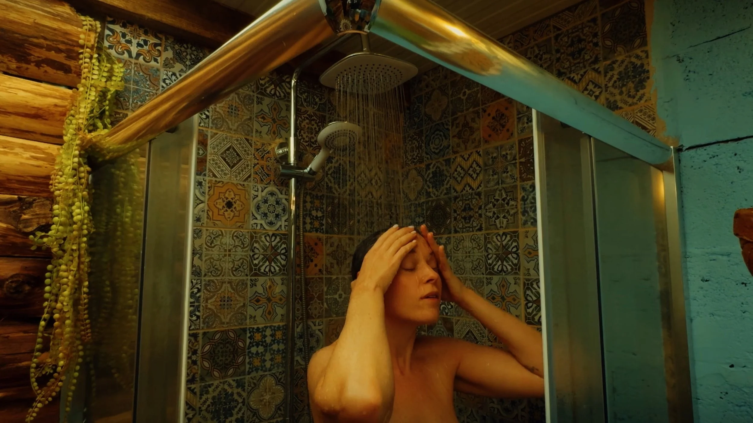 A woman with short dark hair is standing in a shower with her eyes closed, rinsing her hair with water from her hands. The shower has patterned tiles, and there is a glass door and a wooden wall with a hanging plant on the side.