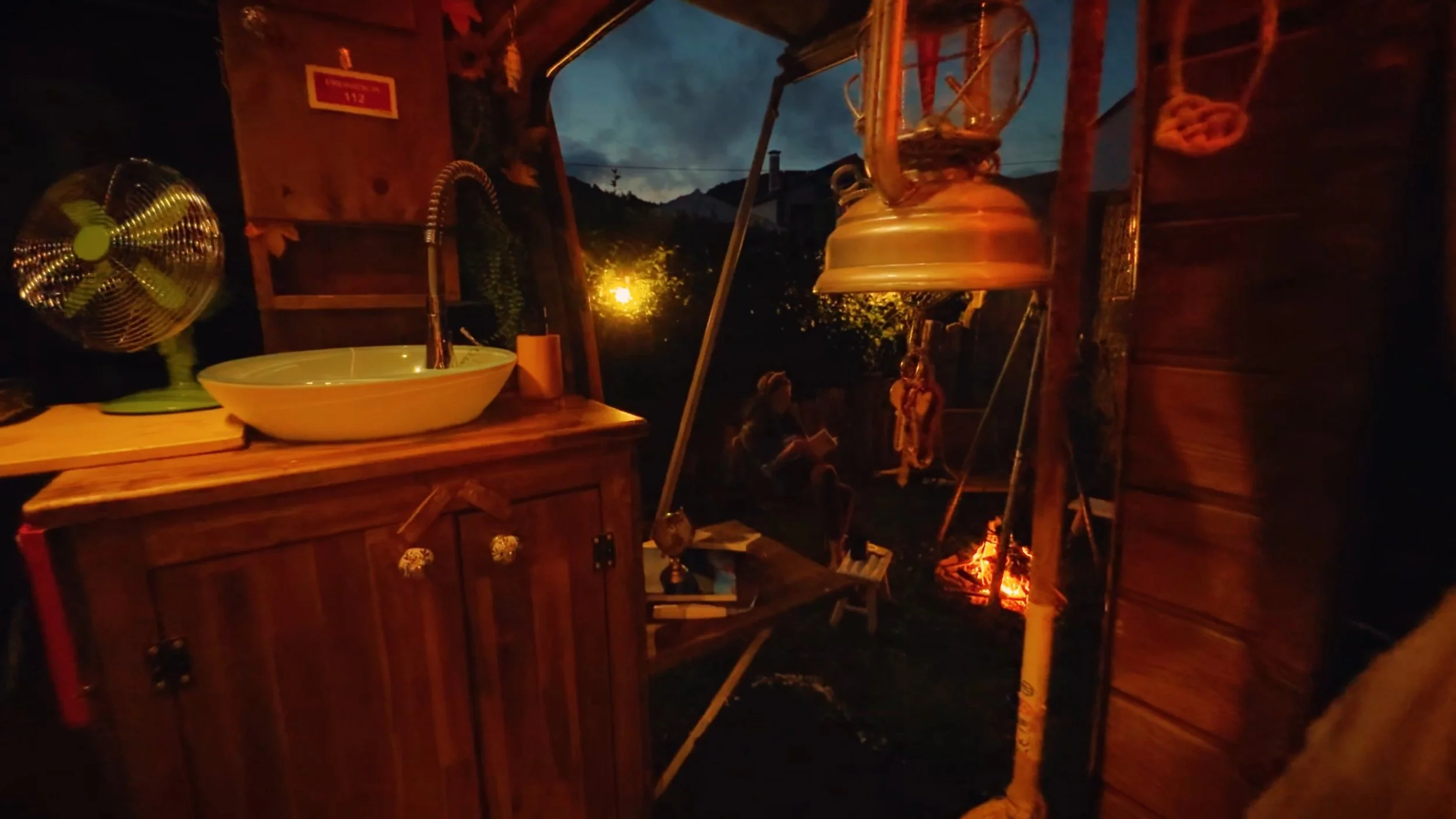 A nighttime outdoor scene with a wooden structure that has a lantern, a fan, and a sink. There is a fire burning in the background with two small figures sitting beside it, and trees and a dark sky are visible in the distance.
