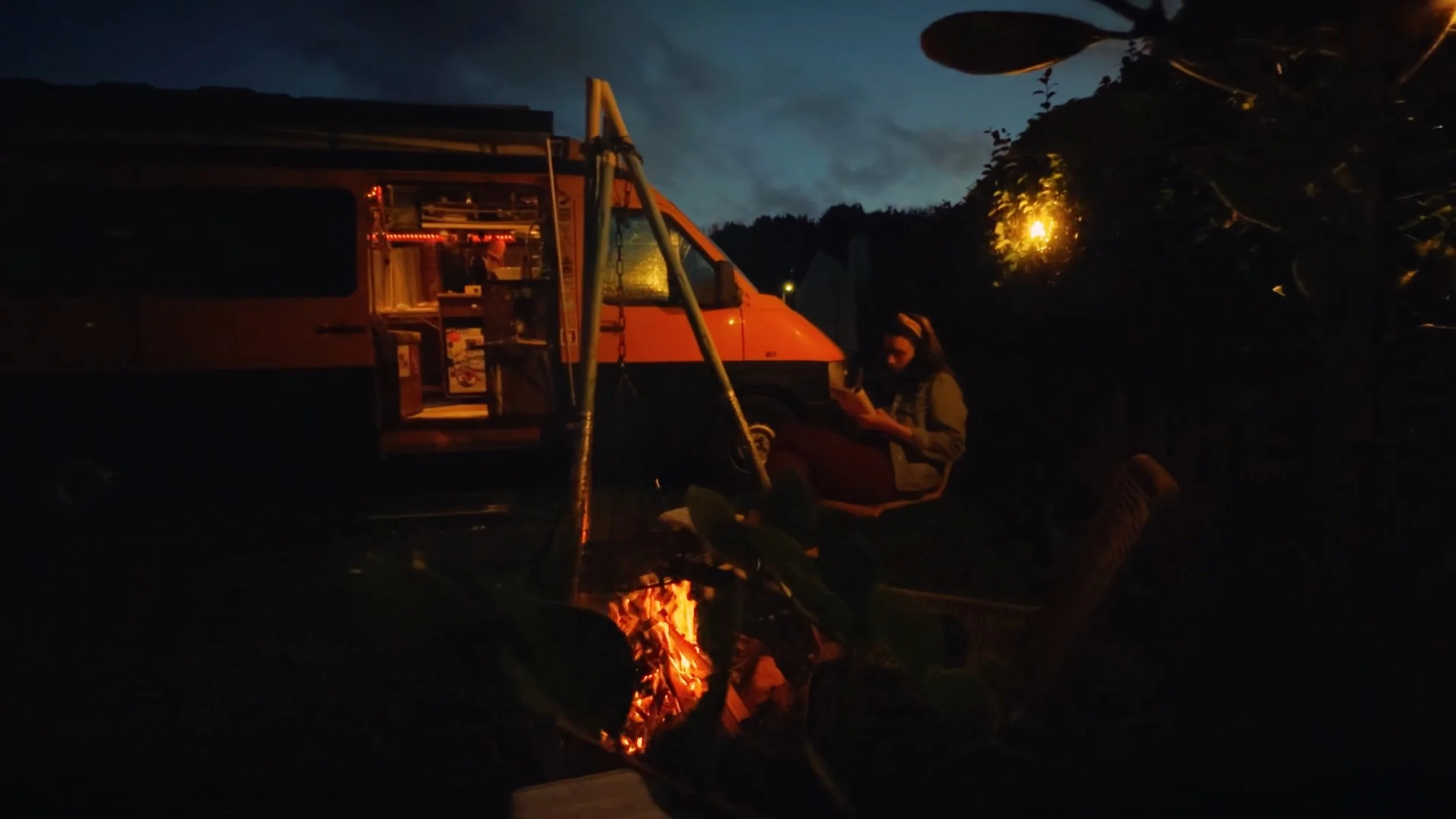 A person sitting on the ground outside near a campfire with a camper van in the background during nighttime. The sky is dark with some clouds and trees are visible.