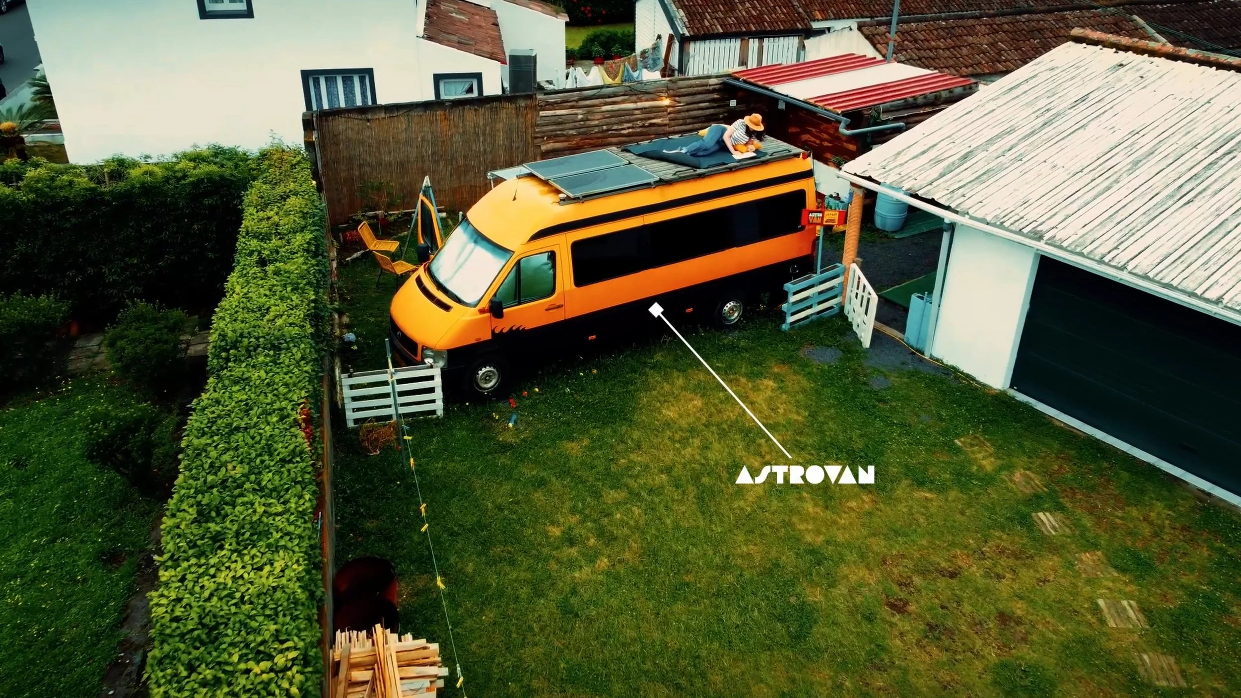 Aerial view of a backyard with a yellow and black camper van, a woman lying on a mattress on top of the van, and a garden with bushes and a pile of firewood. The yard is surrounded by houses with various roof styles.