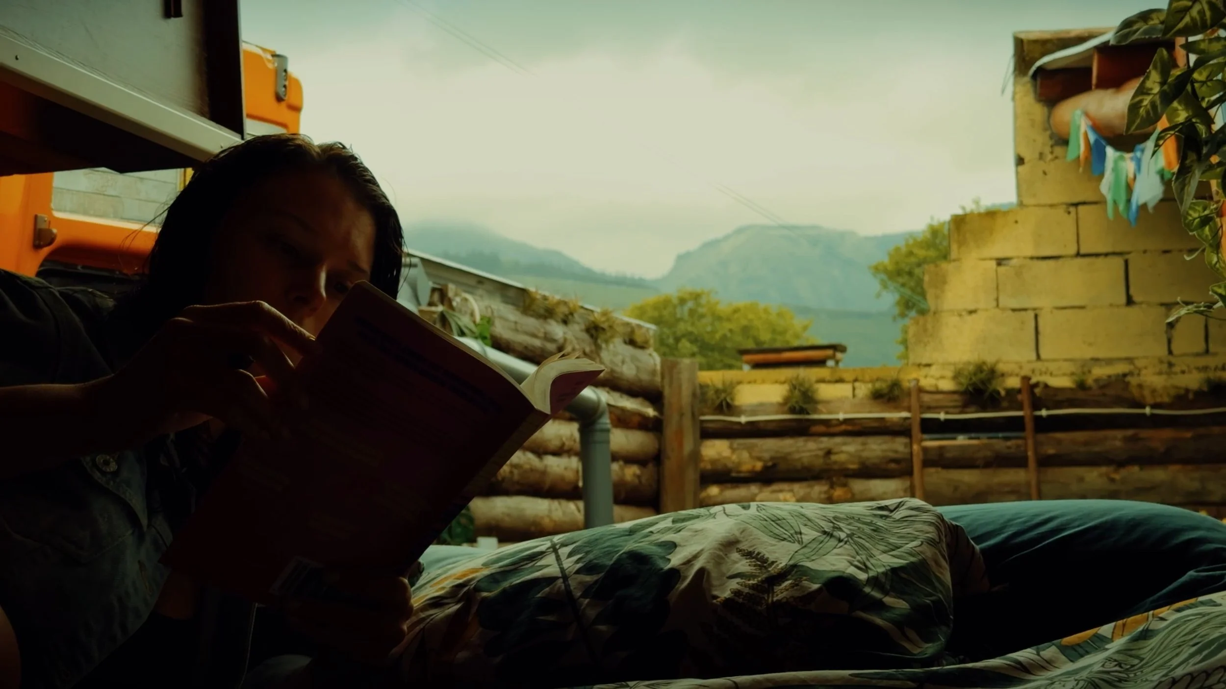 Person with dark hair sitting outdoors, reading a book during daytime, with mountains and a yellow brick wall in the background.