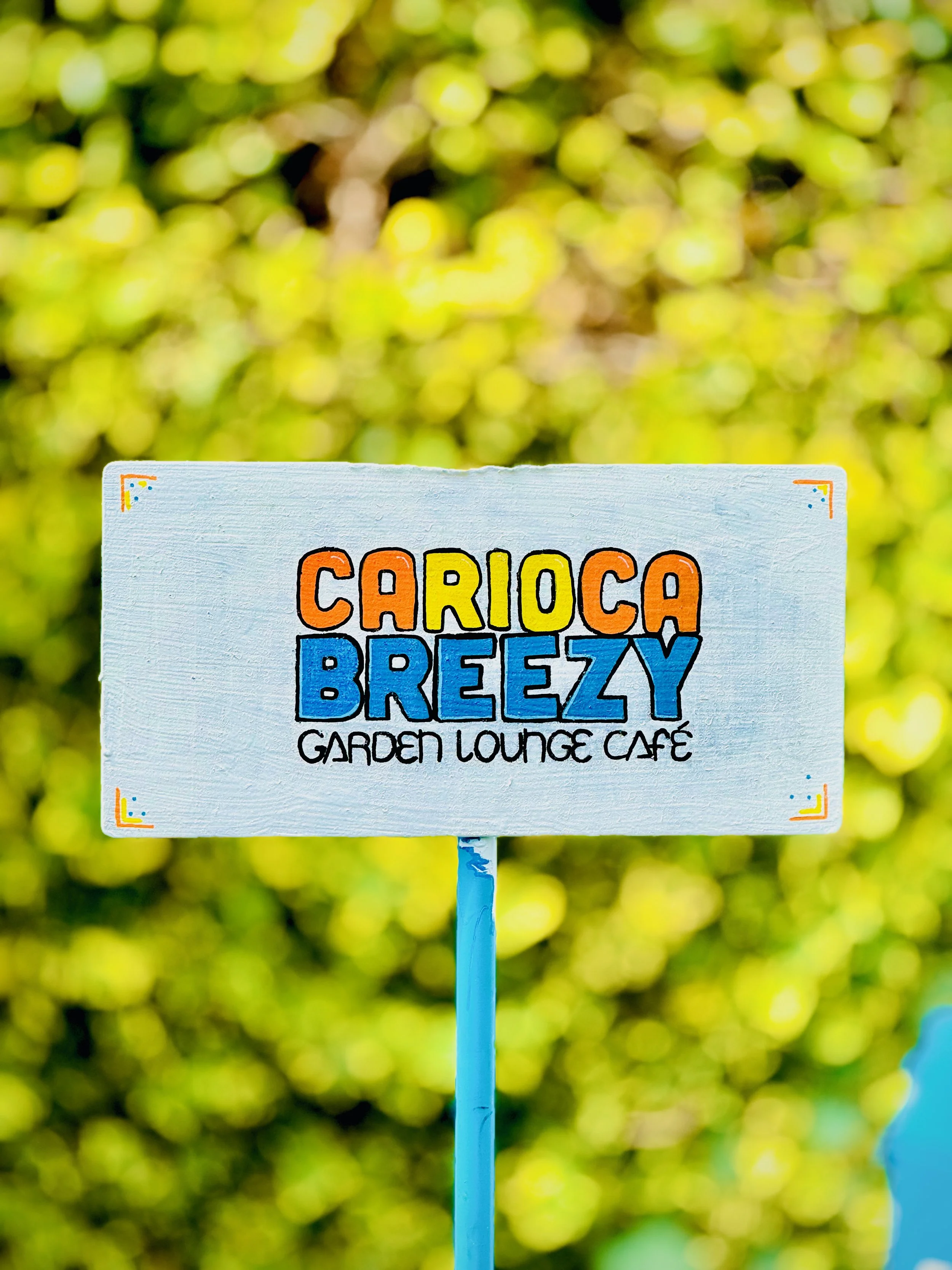 Colorful sign reading 'CARIoca Breezy Garden Lounge Café' with a blurred green background.