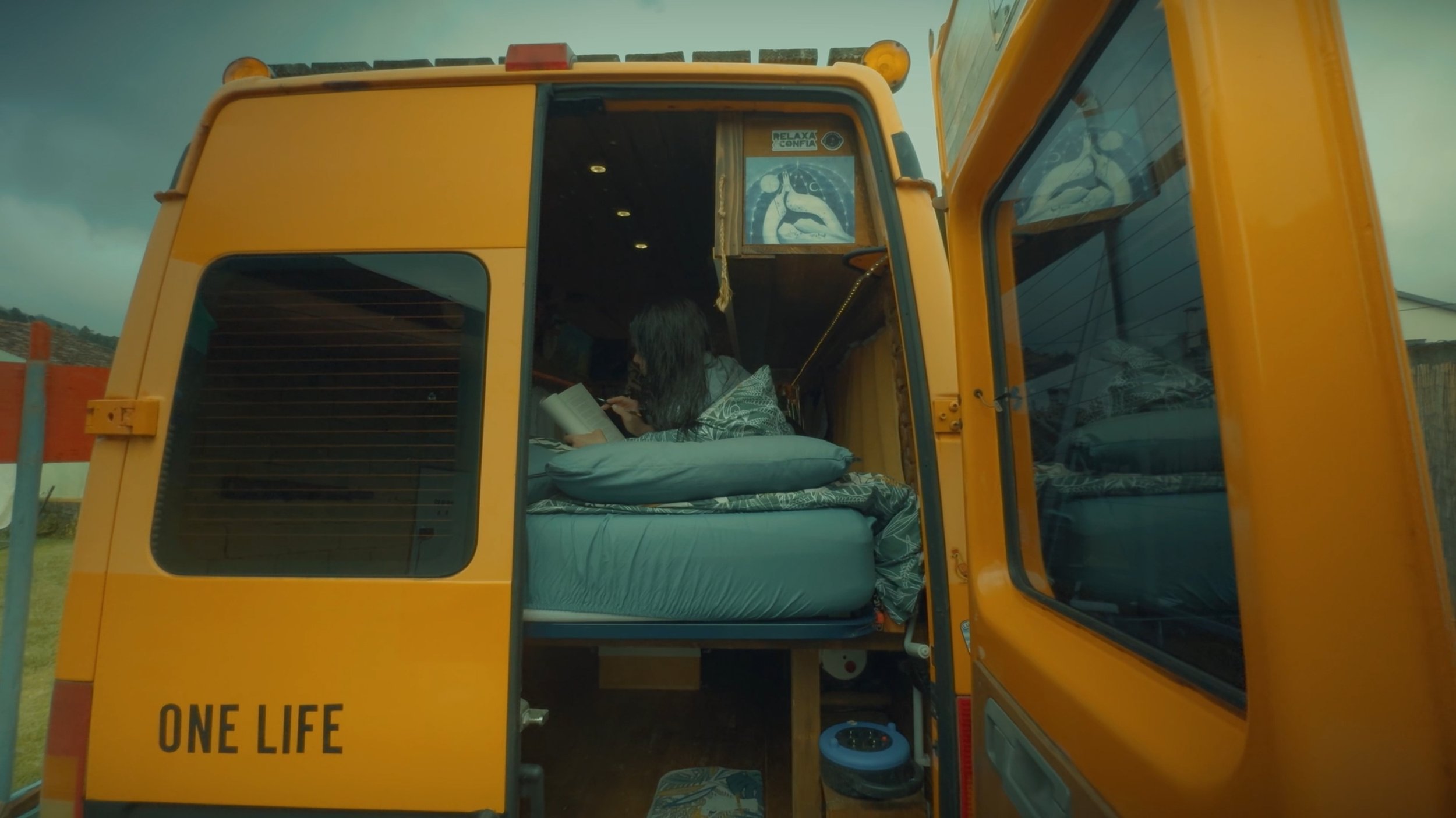 A person with long hair lying in a bed inside a converted yellow school bus, reading a book. The bus has the words 'ONE LIFE' on the back and a window sticker that says 'RELAX & CONFIA.'