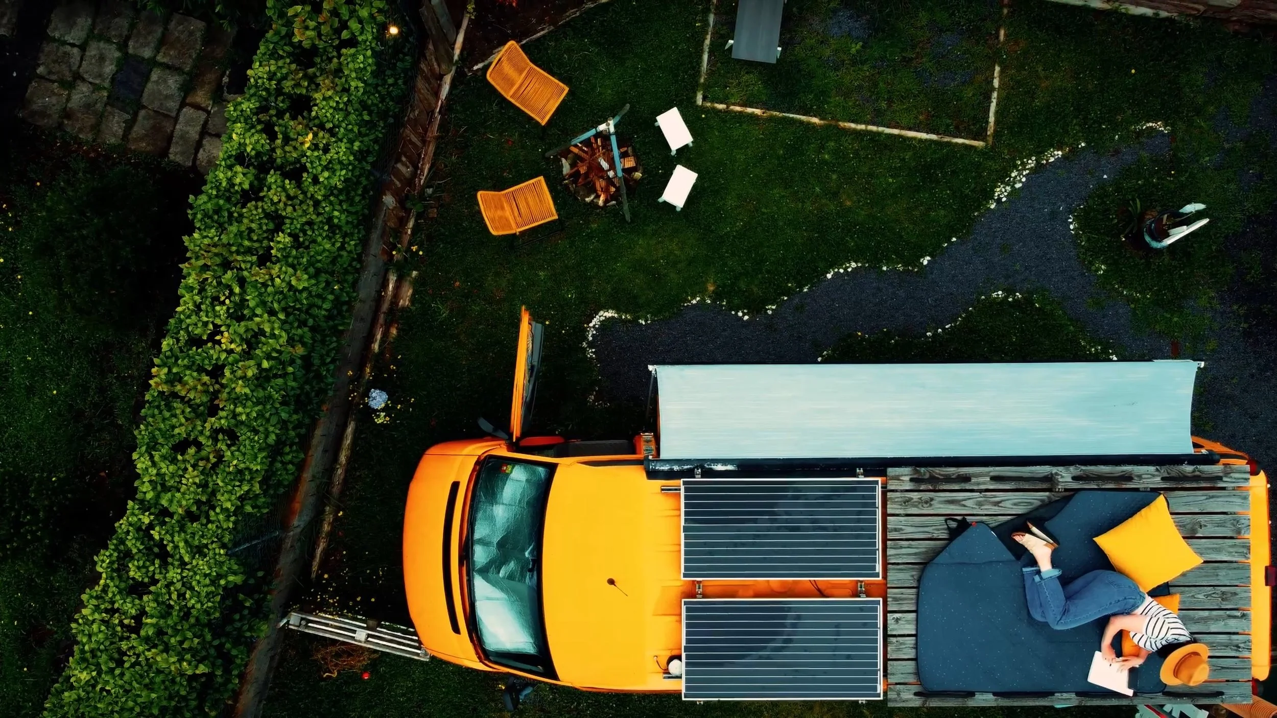 Overhead view of a yard with a yellow solar-powered food truck, outdoor seating, and a person sitting on a yellow cushion reading a book.