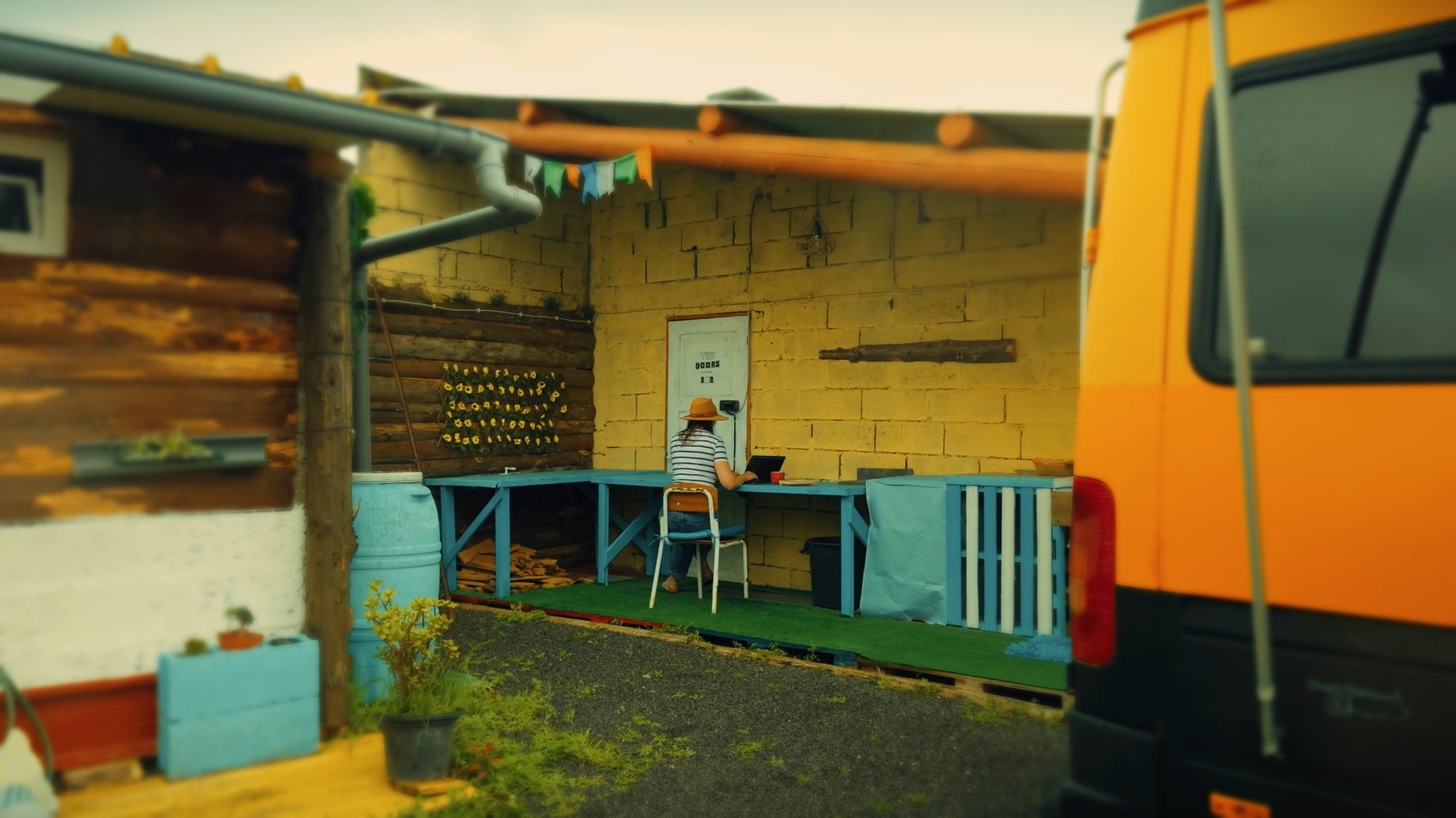 A person with a striped shirt and orange hat sitting at a blue table, working on a laptop in a yellow-walled outdoor space with a blue bench and decorative bunting, seen from behind a yellow vehicle.