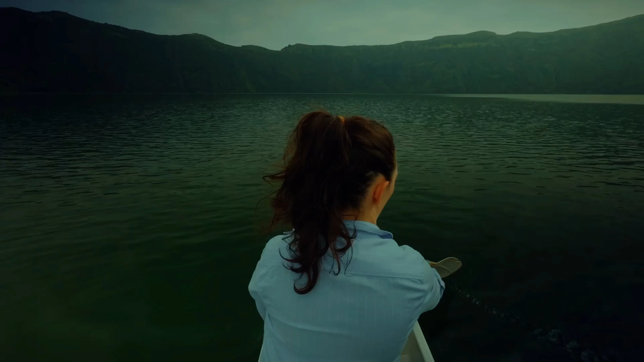 A woman with brown hair tied back, wearing a light blue jacket, is sitting in a boat on a calm body of water surrounded by green mountains, looking towards the horizon.