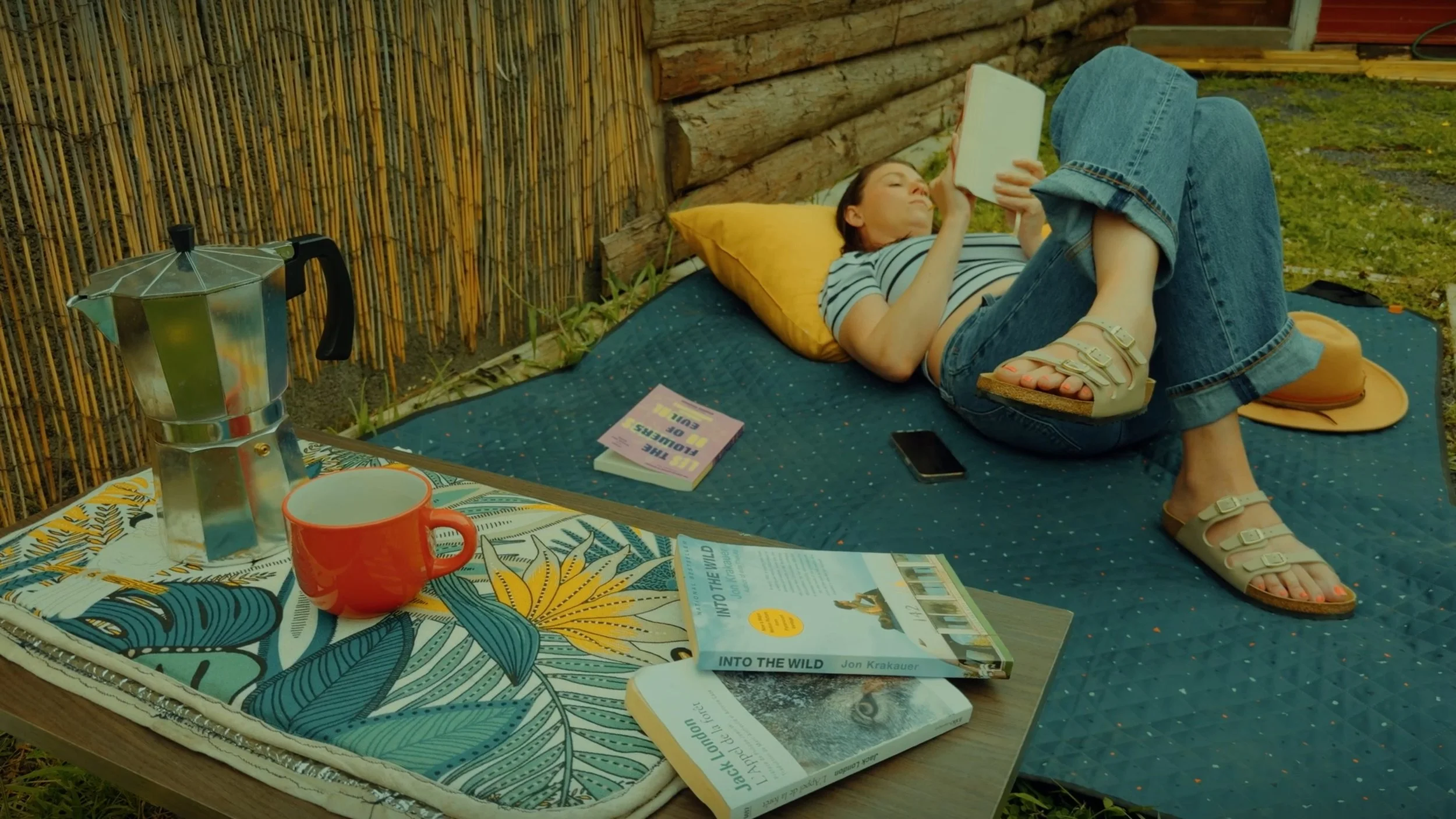 A woman lying on an outdoor bed reading a book in a backyard, with a coffee maker, a red mug, and a magazine titled 'Into the Wild' on the bed. There are other items including a smartphone, a hat, and a tablecloth with a tropical pattern.