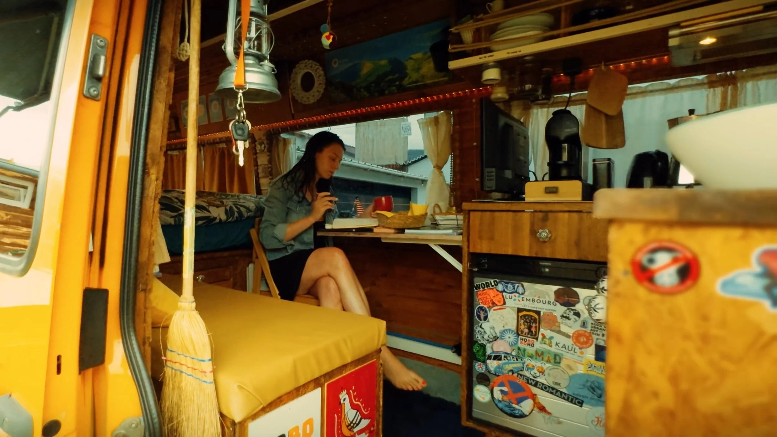 A woman sitting inside a cozy wooden camper van, reading a book and holding a mug, with various decorative items and stickers on the walls and furniture.